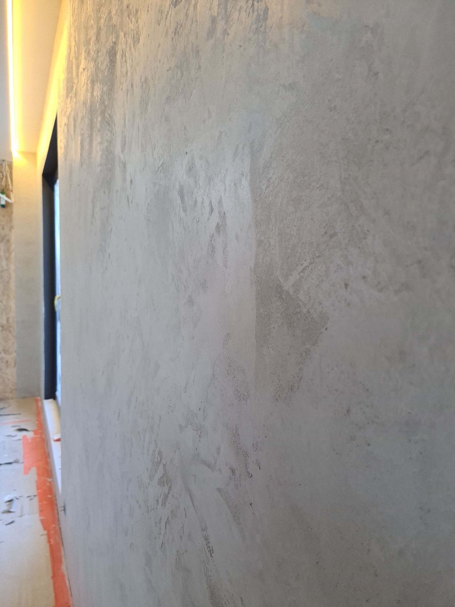 Ściana z dekoracyjną, szarą strukturą, widoczny fragment sufitu z oświetleniem LED oraz fragment drzwi w tle, zabezpieczona podłoga folią malarską.