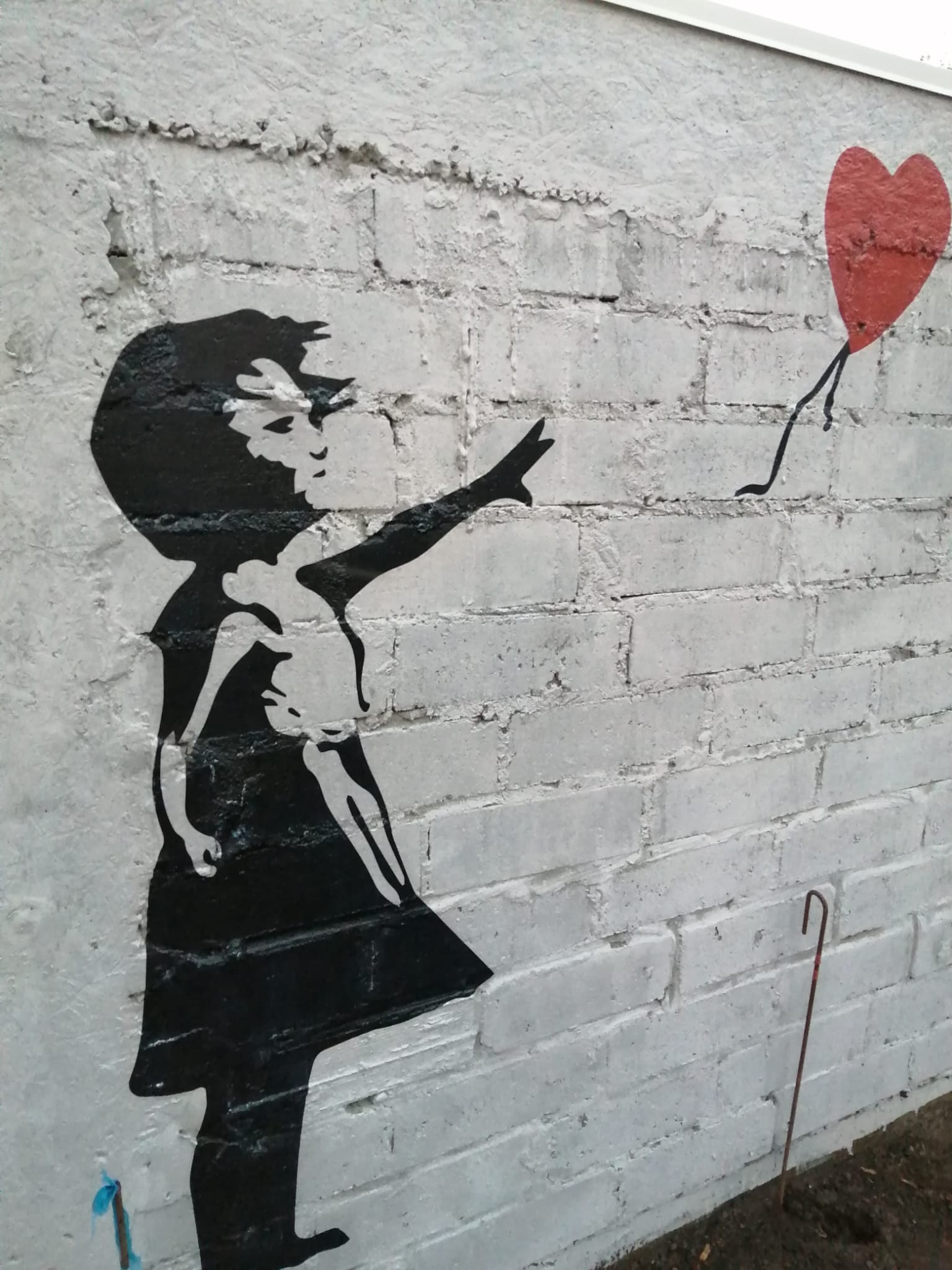Graffiti na białej ceglanej ścianie przedstawia dziewczynkę w czarnej sukience, wyciągającą rękę w stronę czerwonego balonu w kształcie serca na czarnych sznurkach.