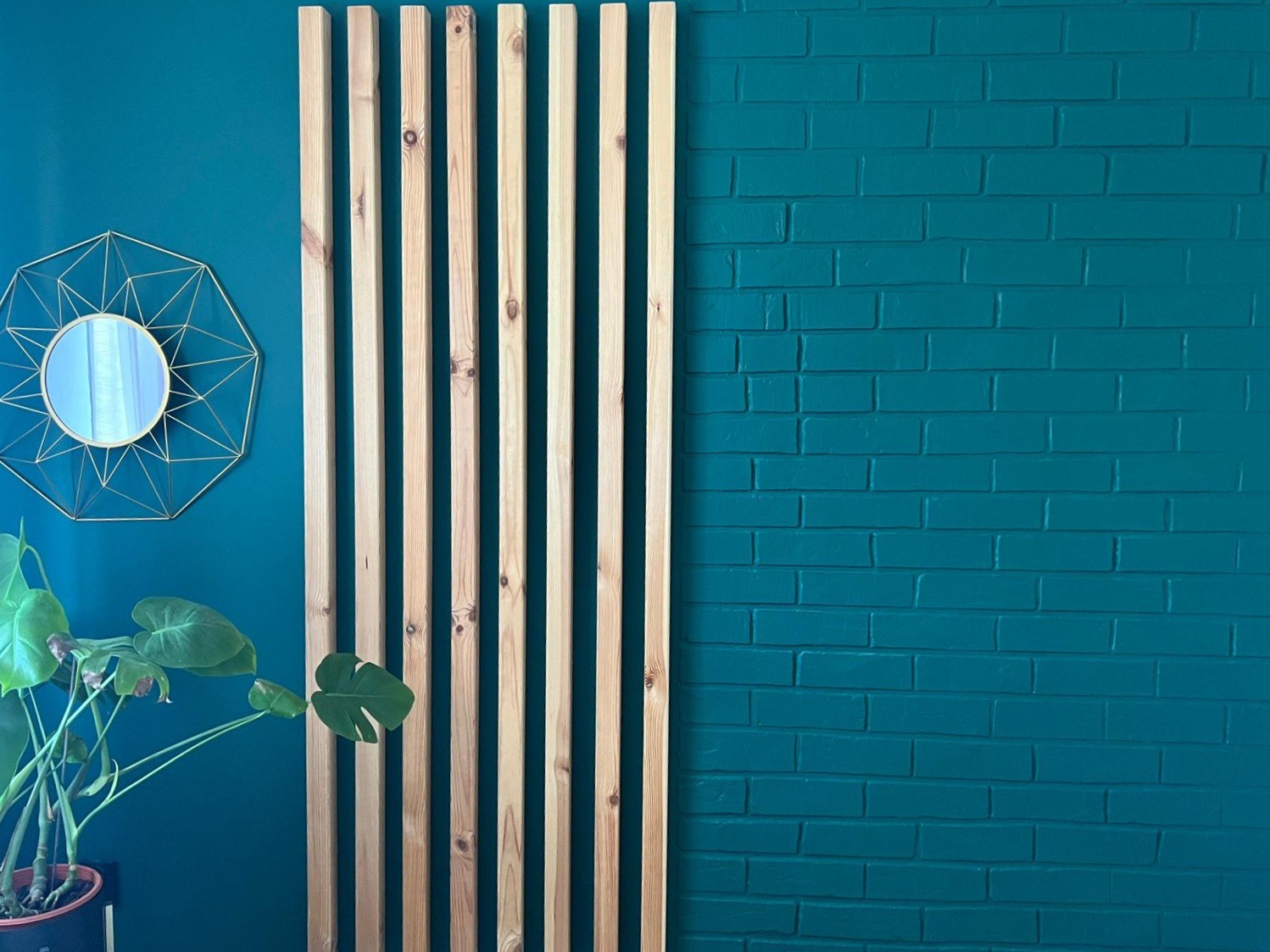 Turkusowa ściana z cegły i drewniany ażur z pionowych listew, okrągłe lustro w złotej ramie i doniczkowa monstera.