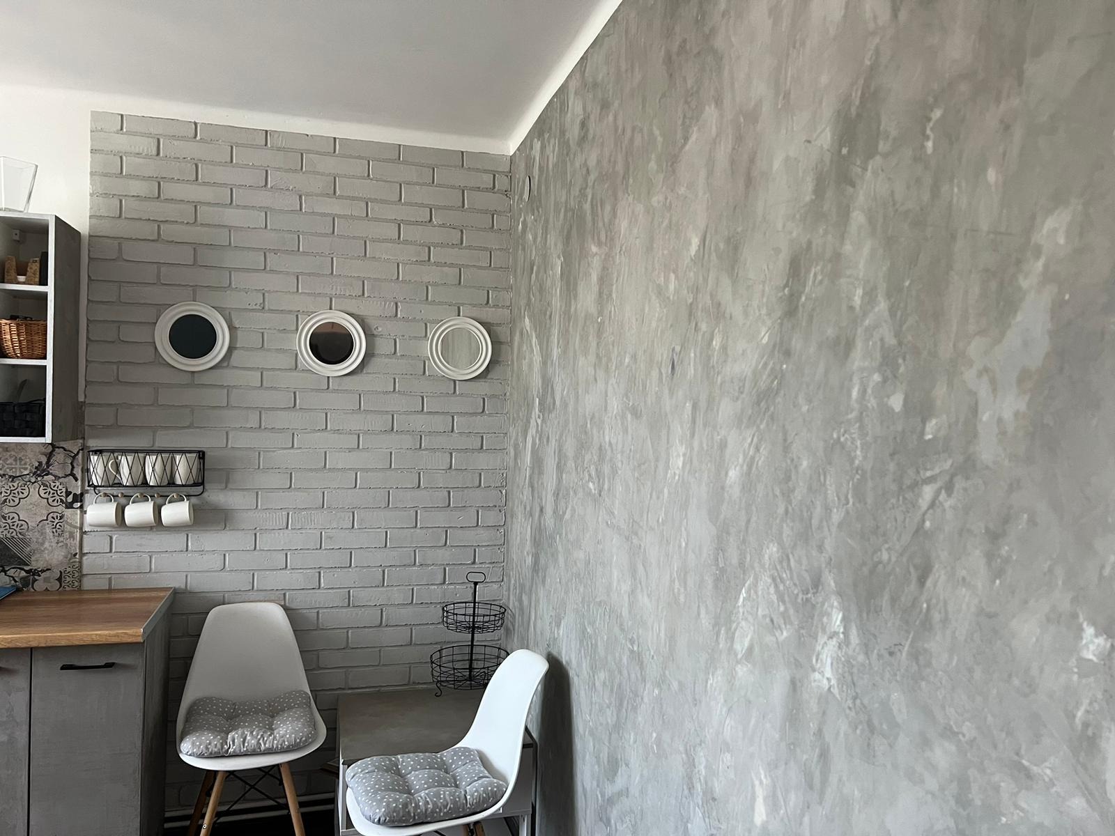 Szara ściana z efektem betonu kontrastuje z białą cegłą, na której wiszą trzy okrągłe lustra, a pod nimi metalowy wieszak na kubki i białe krzesła z poduszkami w szare kropki