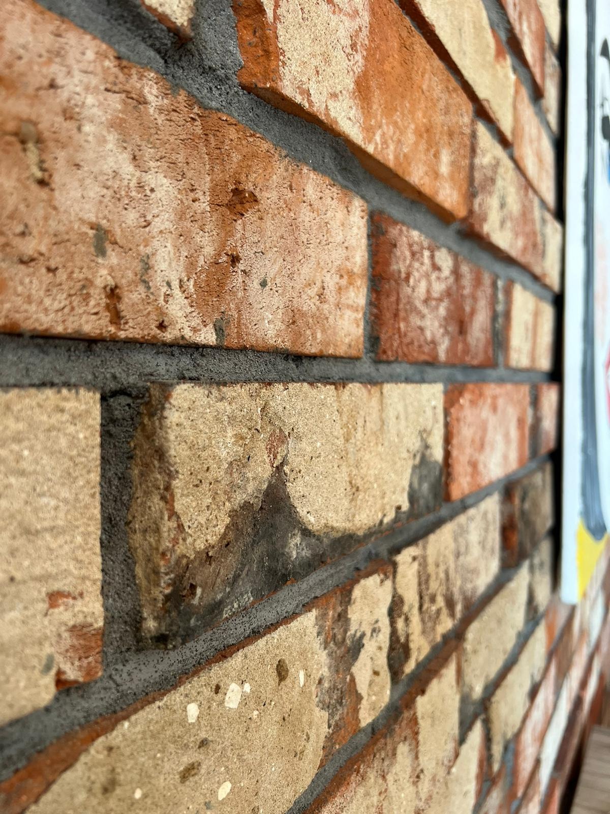 Fragment ściany z cegły klinkierowej w odcieniach beżu i czerwieni, z szarą fugą; widoczne nierówności i tekstura cegieł, ujęcie pod kątem, z boku widoczny fragment obrazu.