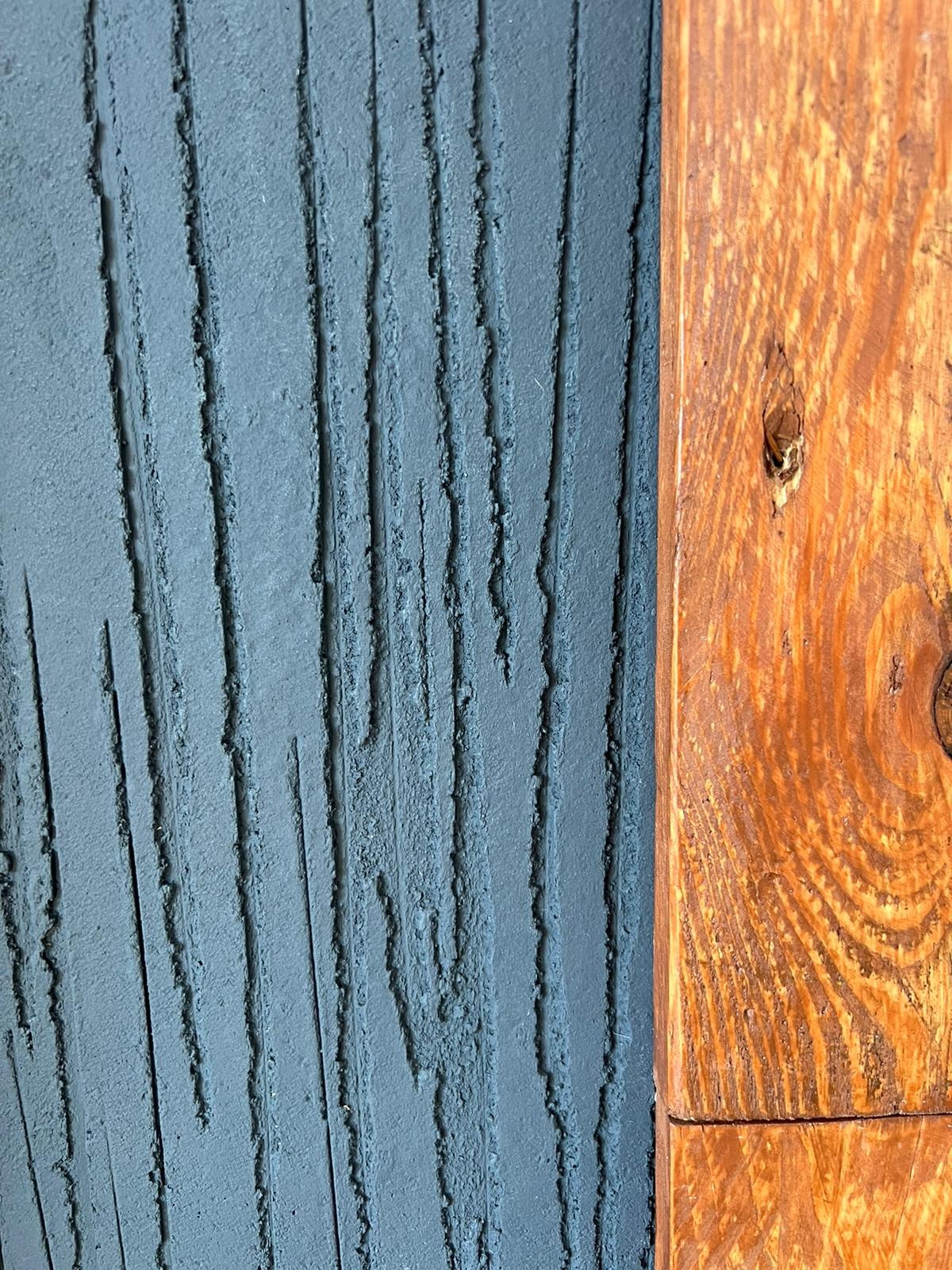 Szara ściana z wyraźną fakturą pionowych rowków sąsiadująca z drewnianą belką o widocznym usłojeniu i sękach.