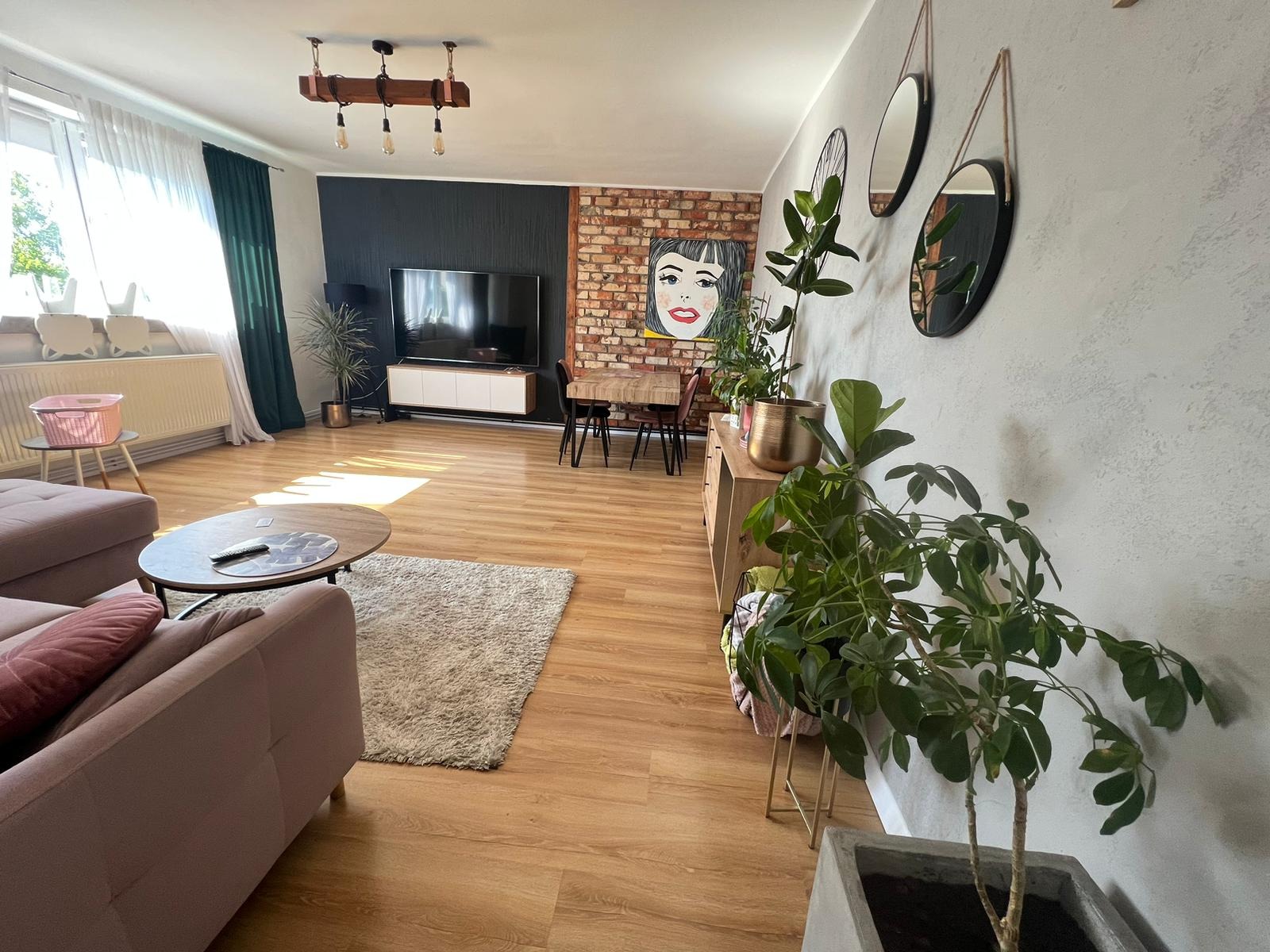 Jasne wnętrze salonu z drewnianą podłogą, ceglaną ścianą z portretem w stylu pop-art, czarną ścianą z telewizorem, szarą sofą i licznymi roślinami doniczkowymi.