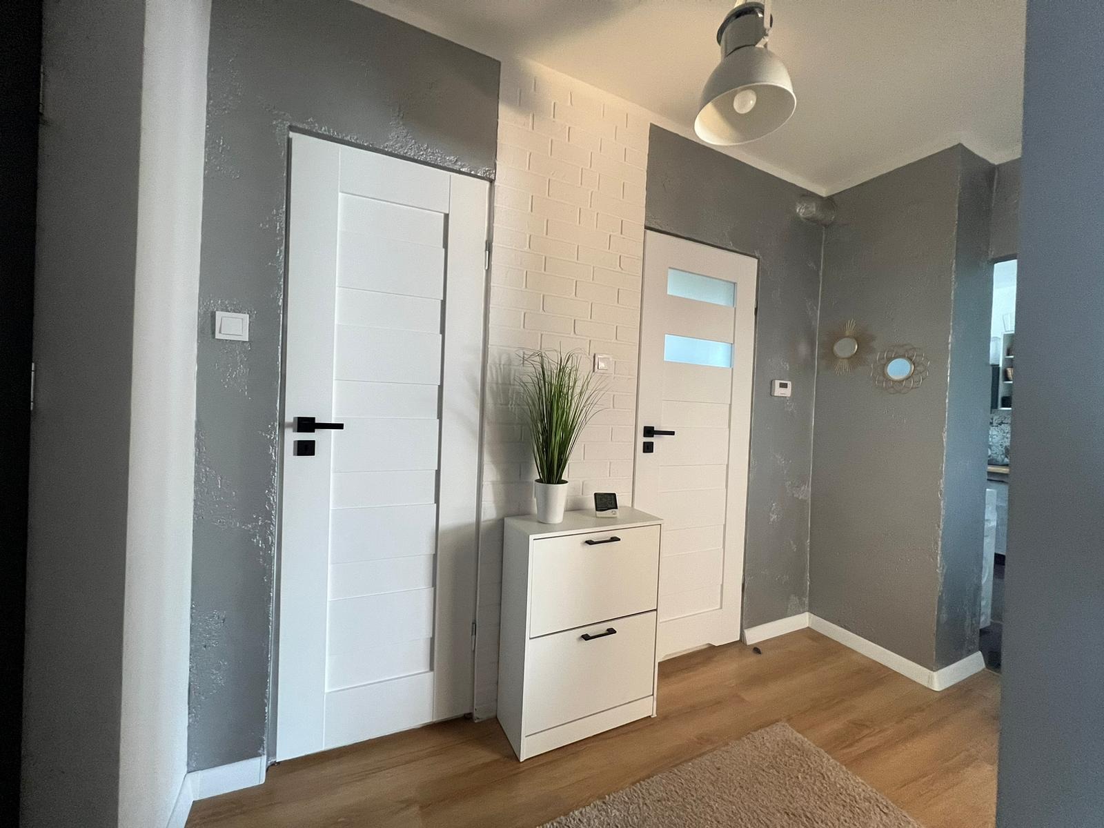Nowoczesny przedpokój z dwiema białymi drzwiami z czarnymi klamkami, szarą ścianą z efektem dekoracyjnym, białą ścianą z imitacją cegły, białą szafką na buty z dekoracyjną trawą i nowoczesną lampą...