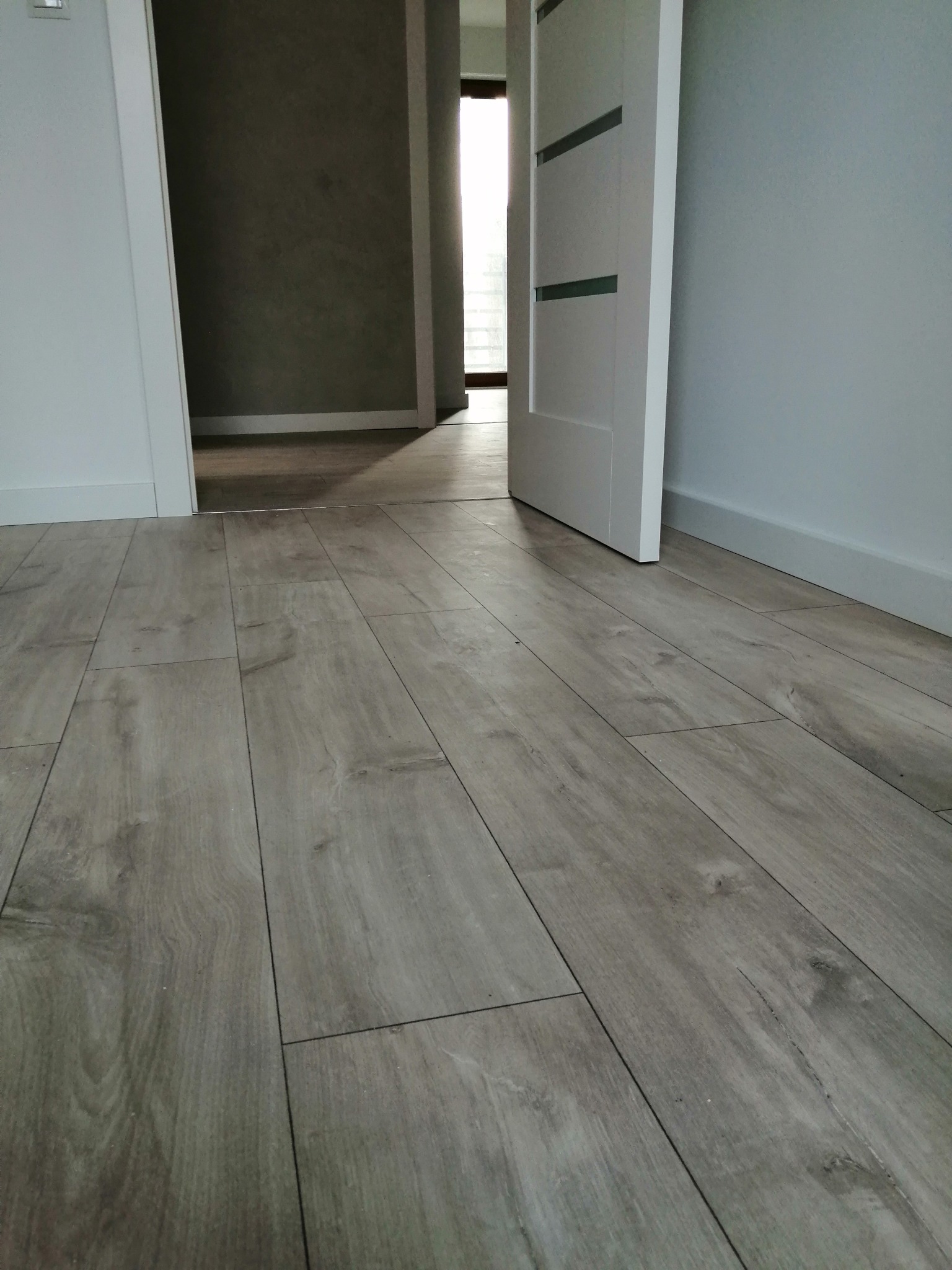 Ułożona podłoga z paneli imitujących jasne drewno, białe listwy przypodłogowe, otwarta biała drzwi wewnętrzne z trzema poziomymi przeszkleniami, szara ściana w tle.