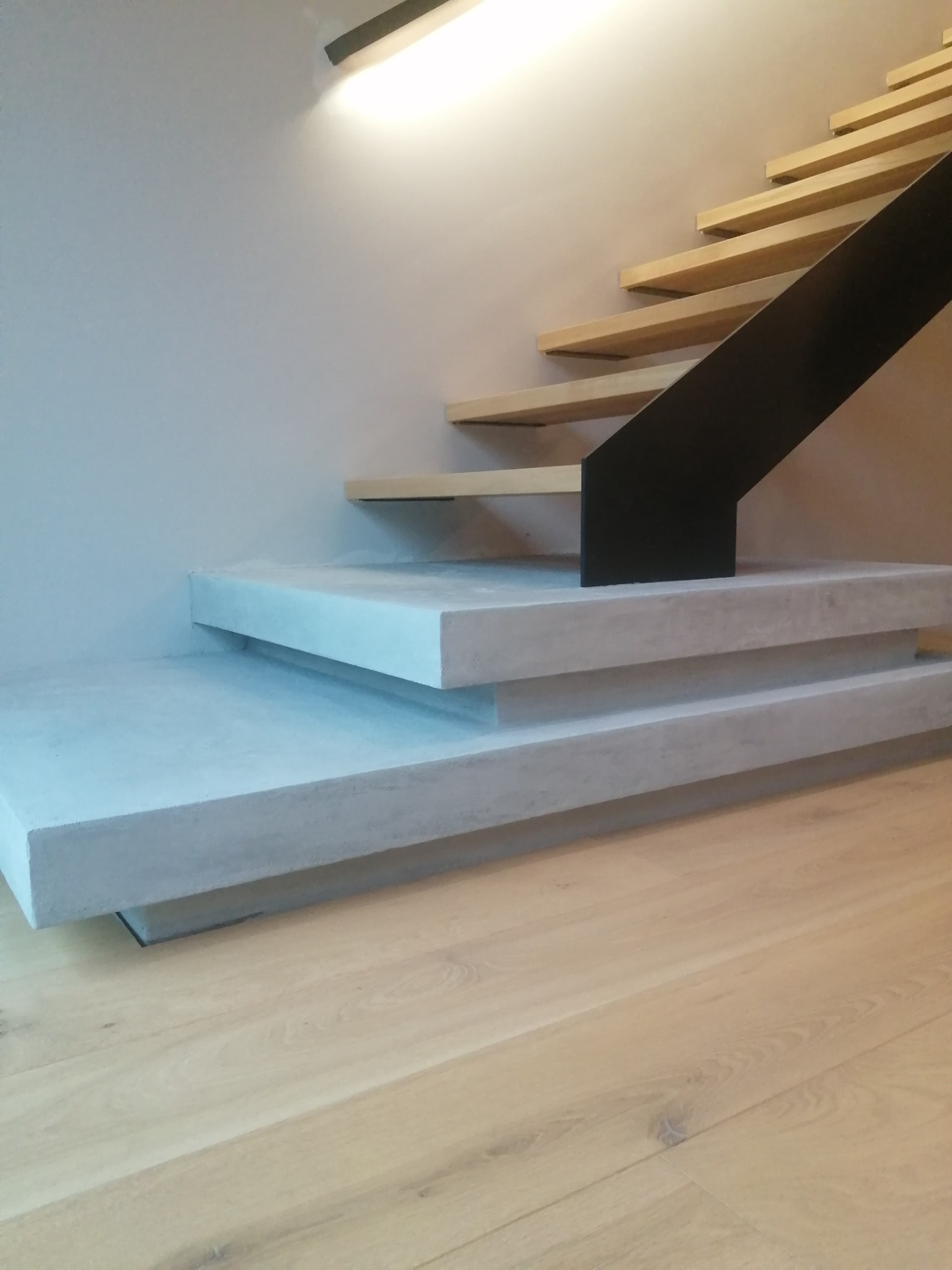 Nowoczesne schody z betonowymi stopniami i drewnianymi podstopnicami, czarna metalowa barierka, oświetlenie punktowe nad schodami, parkiet na podłodze.