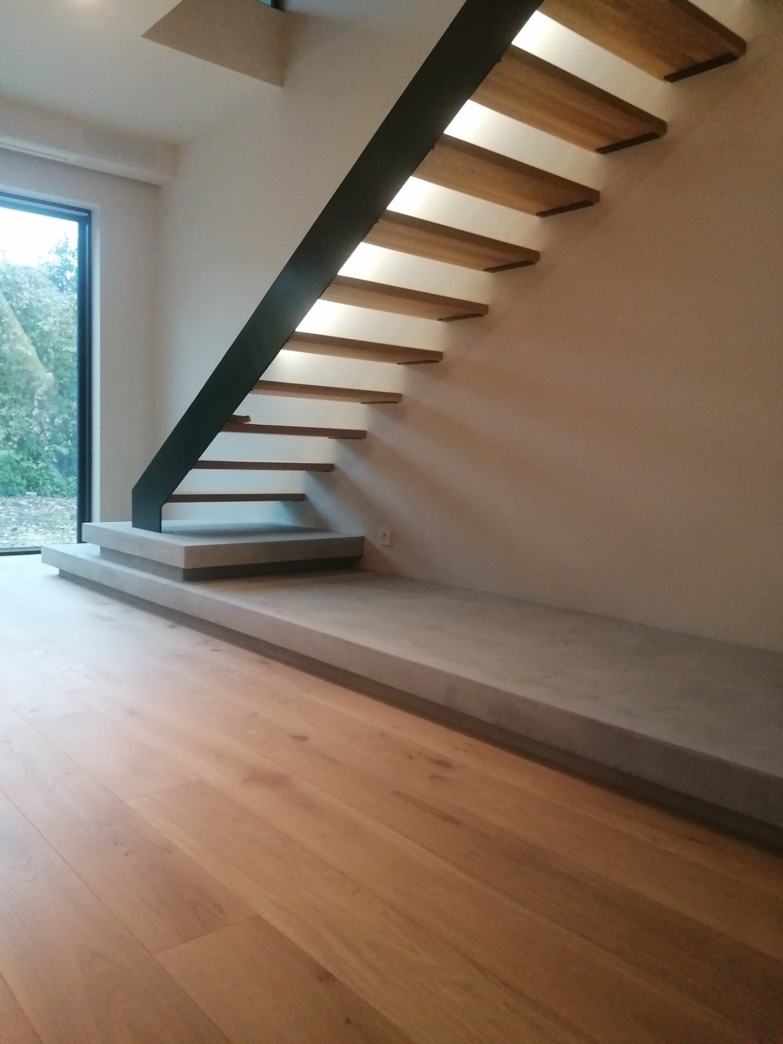 Nowoczesne schody wiszące z drewnianymi stopniami i czarną metalową konstrukcją, oświetlone od spodu, z betonową podstawą i drewnianą podłogą w minimalistycznym wnętrzu.