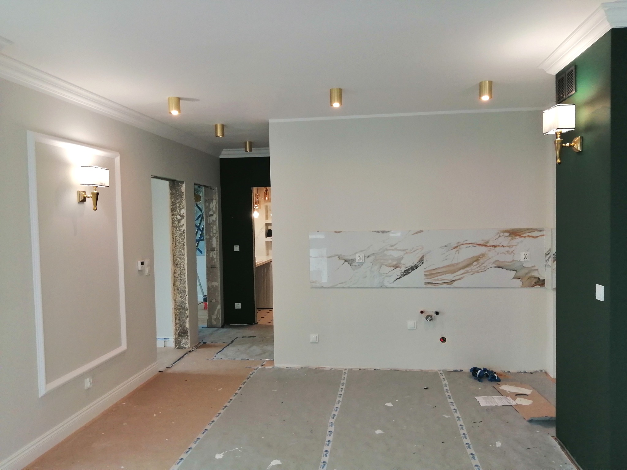 Wnętrze w trakcie prac wykończeniowych: przygotowane podłoże pod posadzkę, fragment ściany z płytą imitującą marmur, zainstalowane punkty oświetleniowe i ozdobne listwy przysufitowe.