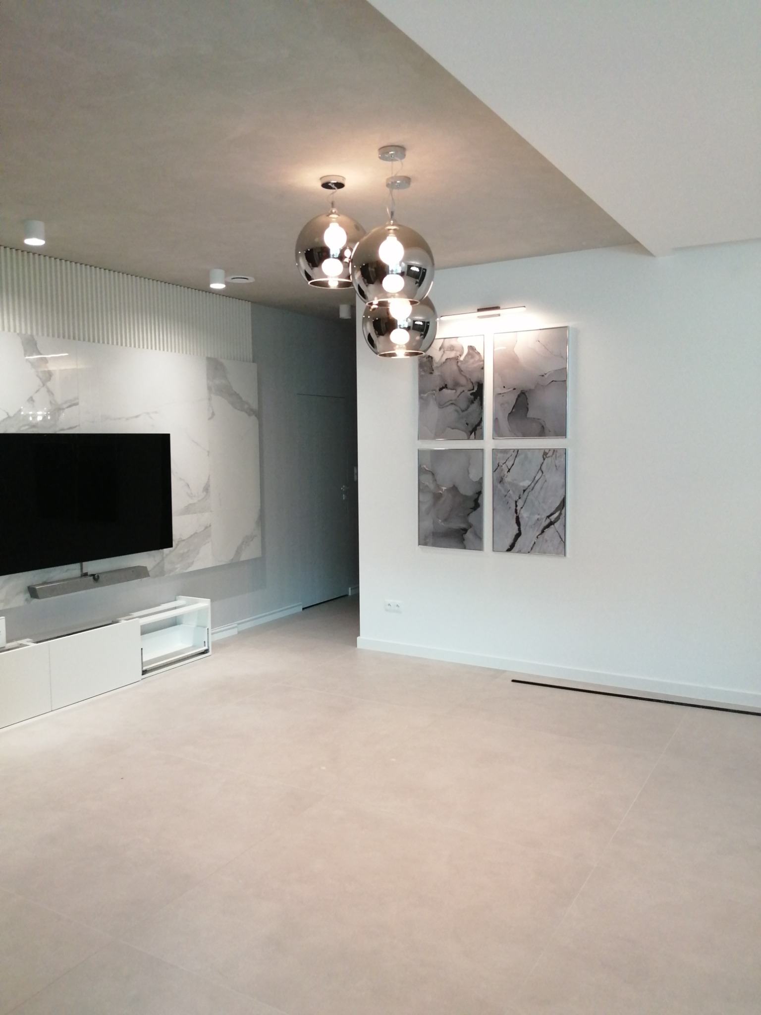 Nowoczesny salon z białymi ścianami, szarą podłogą z płytek, dekoracyjną ścianą z marmurowym wzorem, trzema okrągłymi lampami wiszącymi i czterema obrazami w szarych tonacjach.