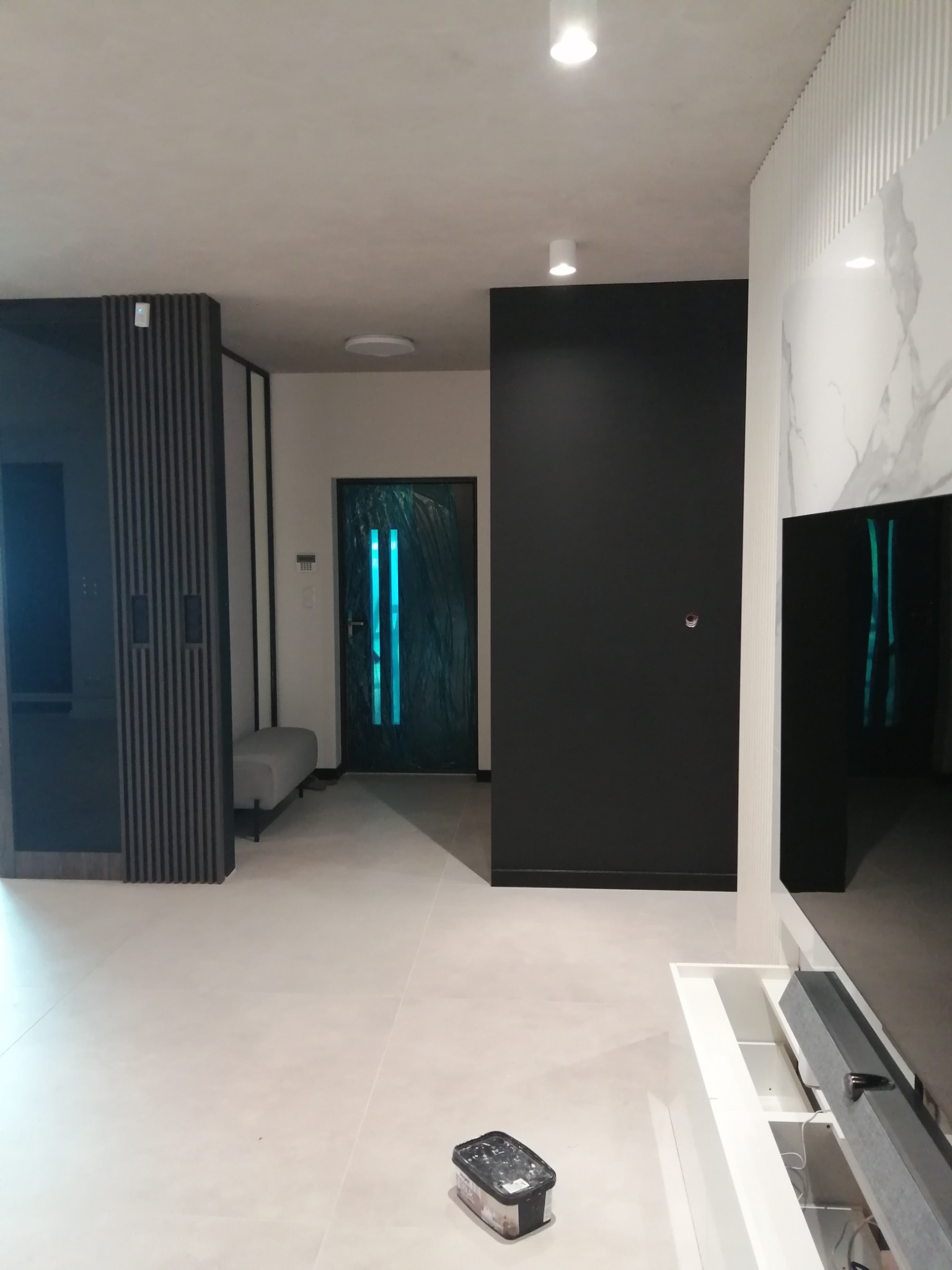 Nowoczesne wnętrze mieszkania po remoncie: jasna podłoga, czarne ściany, dekoracyjny panel ścienny z efektem marmuru, drzwi zabezpieczone folią ochronną, widoczny pojemnik z narzędziami na podłodze.