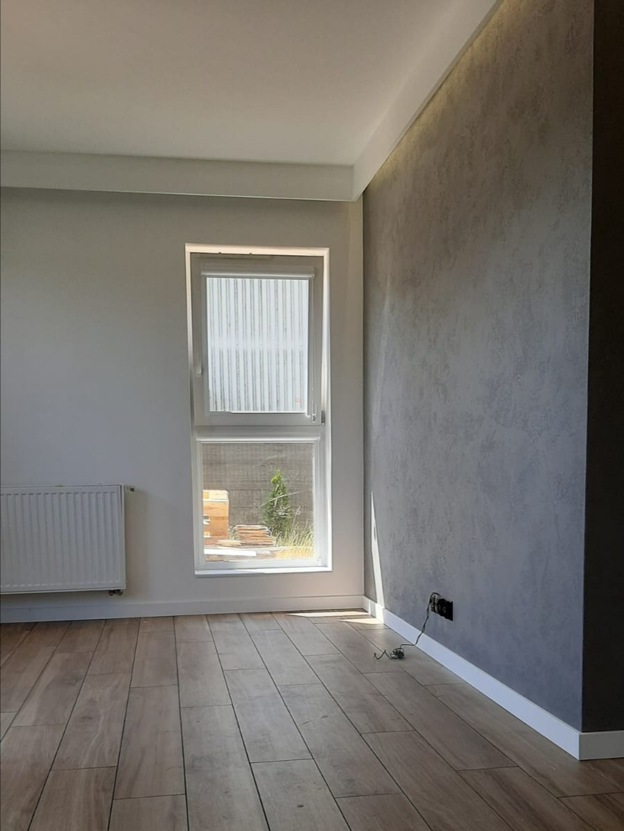 Pomalowane na szaro pomieszczenie z drewnianą podłogą, białym grzejnikiem i oknem z widokiem na zewnątrz, z detalami takimi jak listwy przypodłogowe i gniazdko elektryczne.