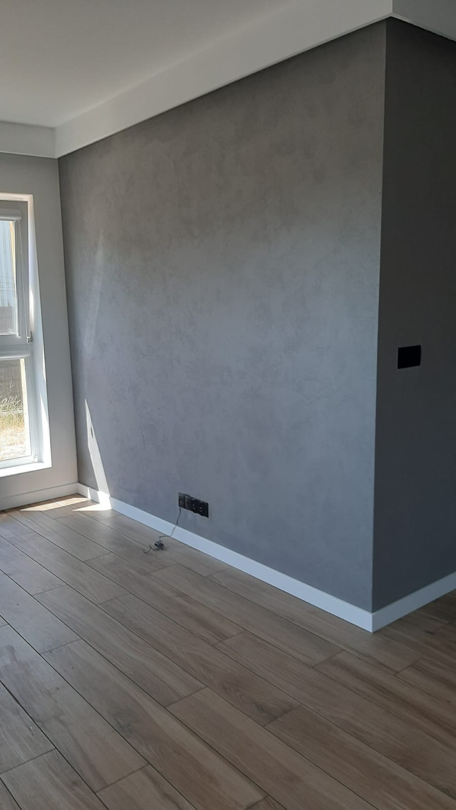 Szara ściana z efektem dekoracyjnym, listwa przypodłogowa, drewnopodobna podłoga i okno z widocznym światłem słonecznym w rogu pokoju.