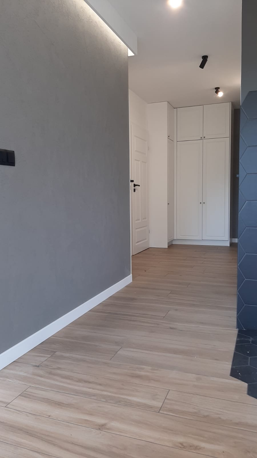 Szara ściana z oświetleniem LED przy suficie, drewnopodobna podłoga, białe drzwi i szafa w tle oraz fragment ściany wykończonej heksagonalnymi płytkami.