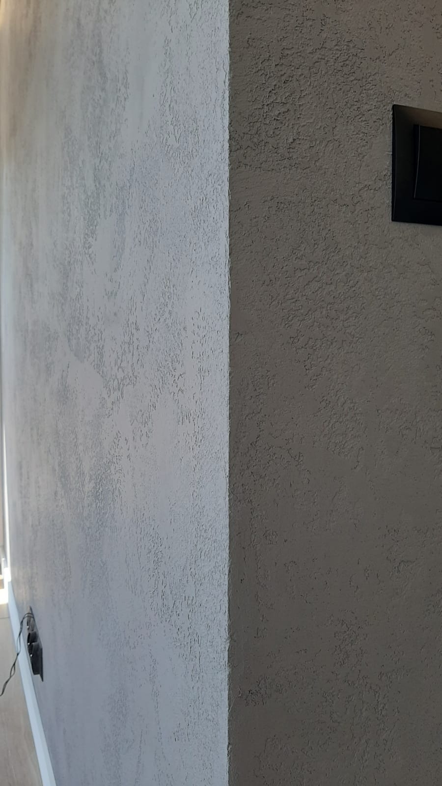 Szara ściana z efektem dekoracyjnym typu baranek, widoczny narożnik i gniazdko elektryczne z czarną ramką.