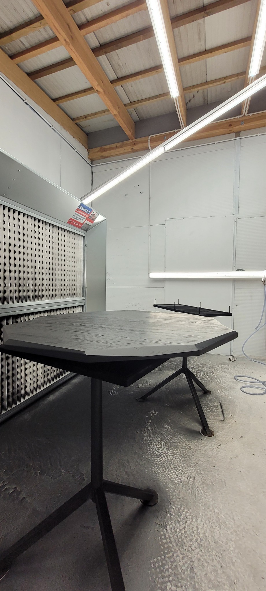 Ośmiokątny stół z drewna po lakierowaniu w kabinie lakierniczej z widocznymi filtrami powietrza.