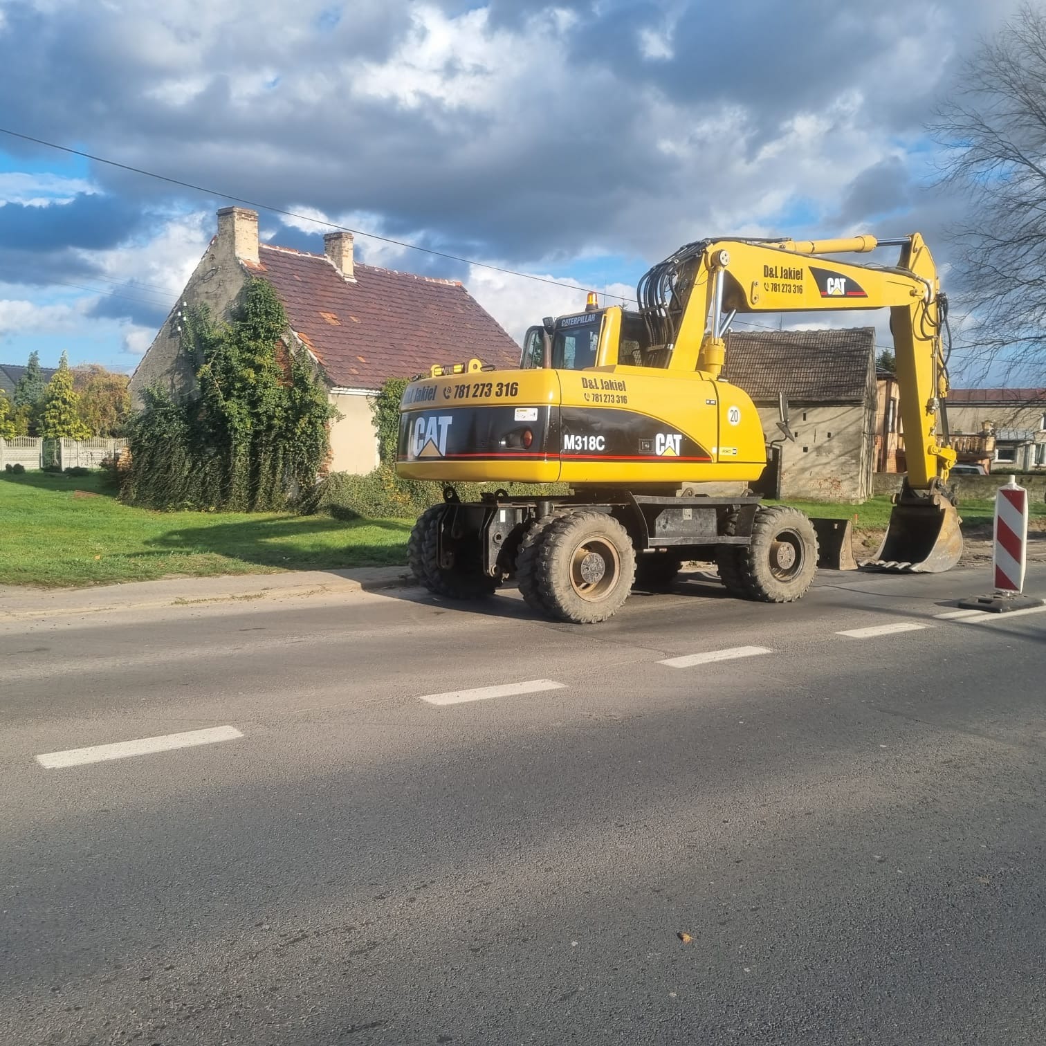 Żółta koparka kołowa CAT M318C firmy D&L Jakiel stoi na asfaltowej drodze z białymi pasami, w tle widoczny dom z ceglanym dachem i fragment trawnika.
