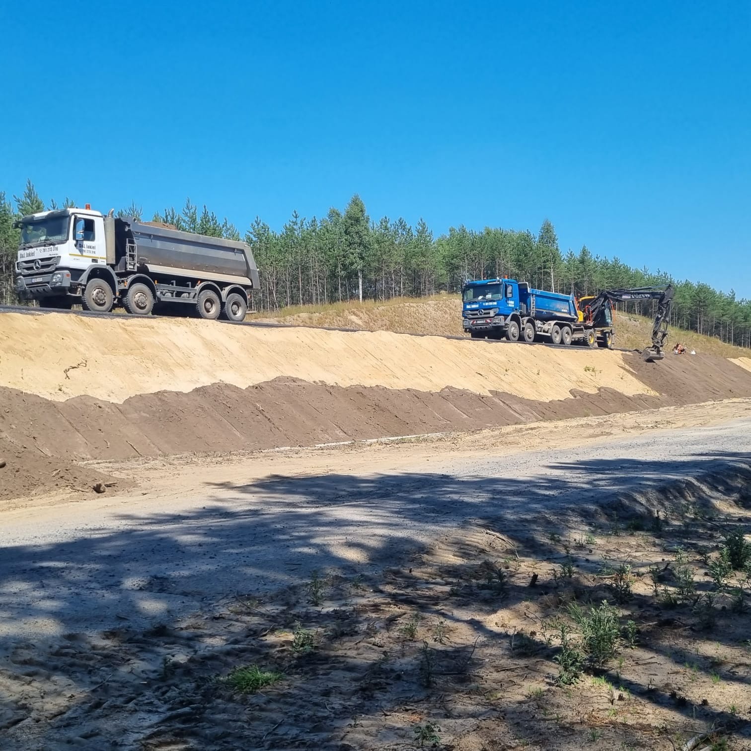 Dwa samochody ciężarowe i koparka na placu budowy, świeżo usypana ziemia w tle, las w oddali, błękitne niebo.