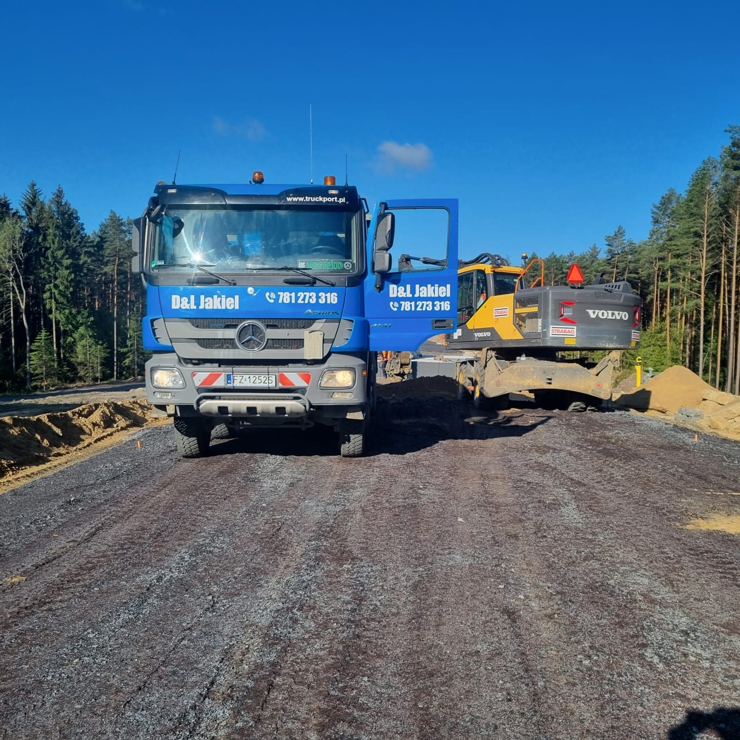 Niebieska ciężarówka Mercedes z otwartymi drzwiami i numerem telefonu firmy D&L Jakiel, obok żółtej koparki Volvo na nowo utwardzonej drodze żwirowej, w tle las iglasty i błękitne niebo.