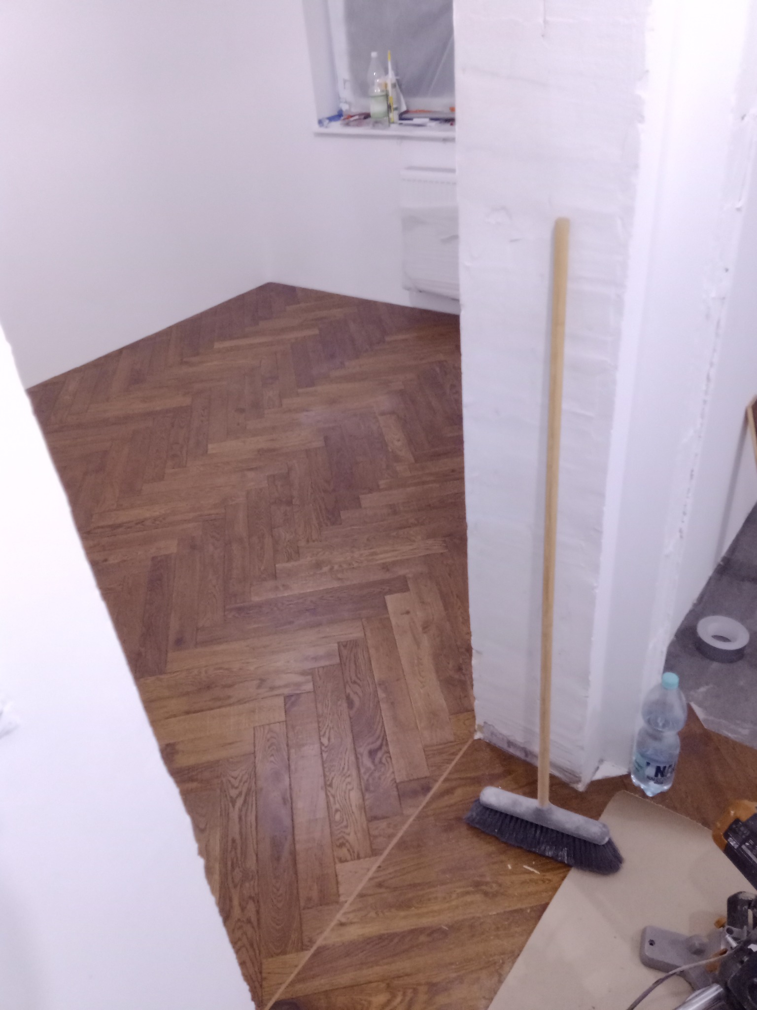 Ułożony parkiet w jodełkę w rogu pokoju, widoczna szczotka oparta o ścianę, butelka wody i fragment piły ukosowej, niedokończone wykończenie ściany.
