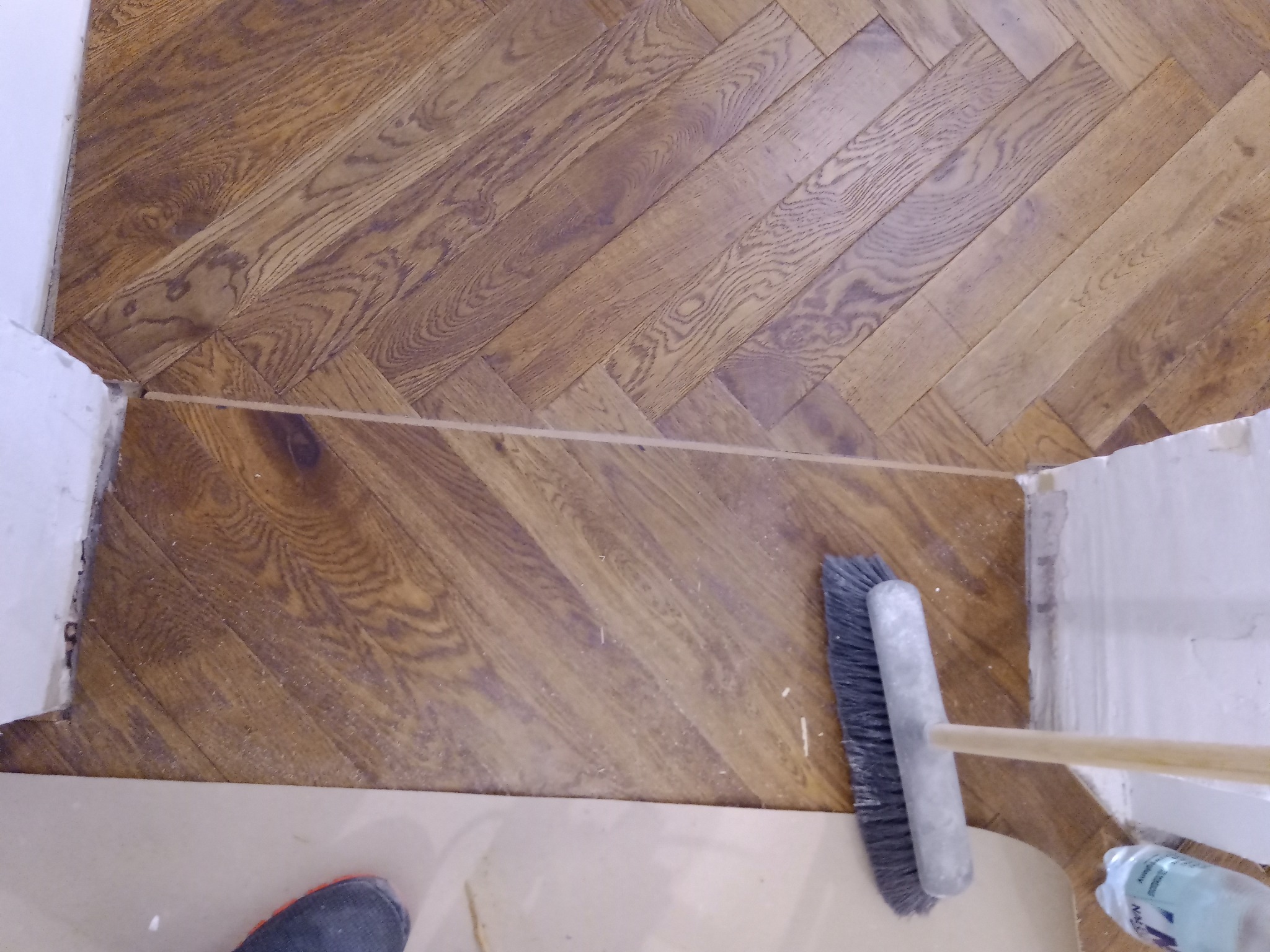 Parkiet ułożony w jodełkę, szczotka do zamiatania i widoczne drobne ślady po cięciu przy ścianie.