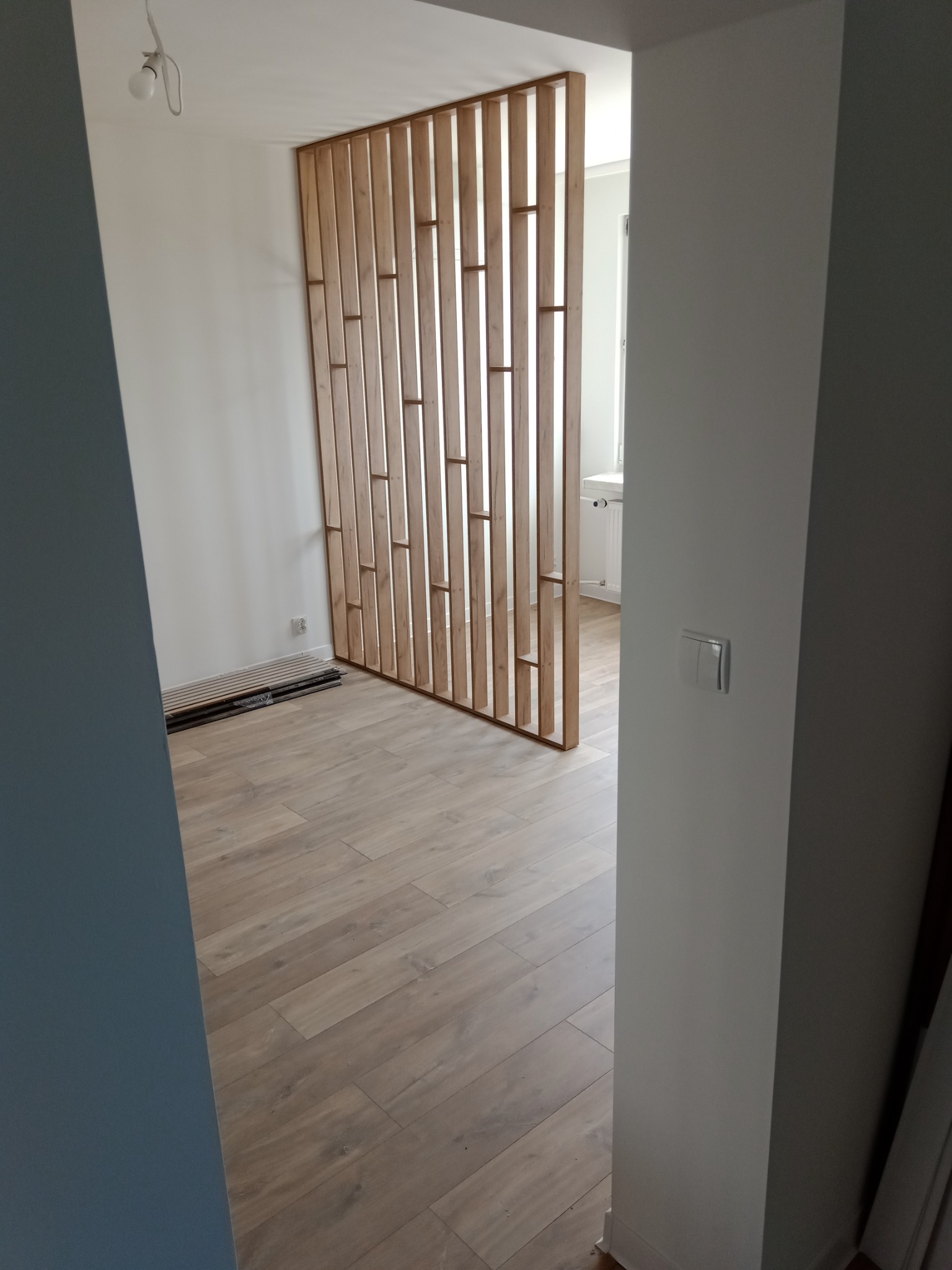 Drewniana ażurowa ścianka działowa z pionowych listew, widoczna z perspektywy wejścia do pomieszczenia, na podłodze panele, ściany pomalowane na biało i szaro.