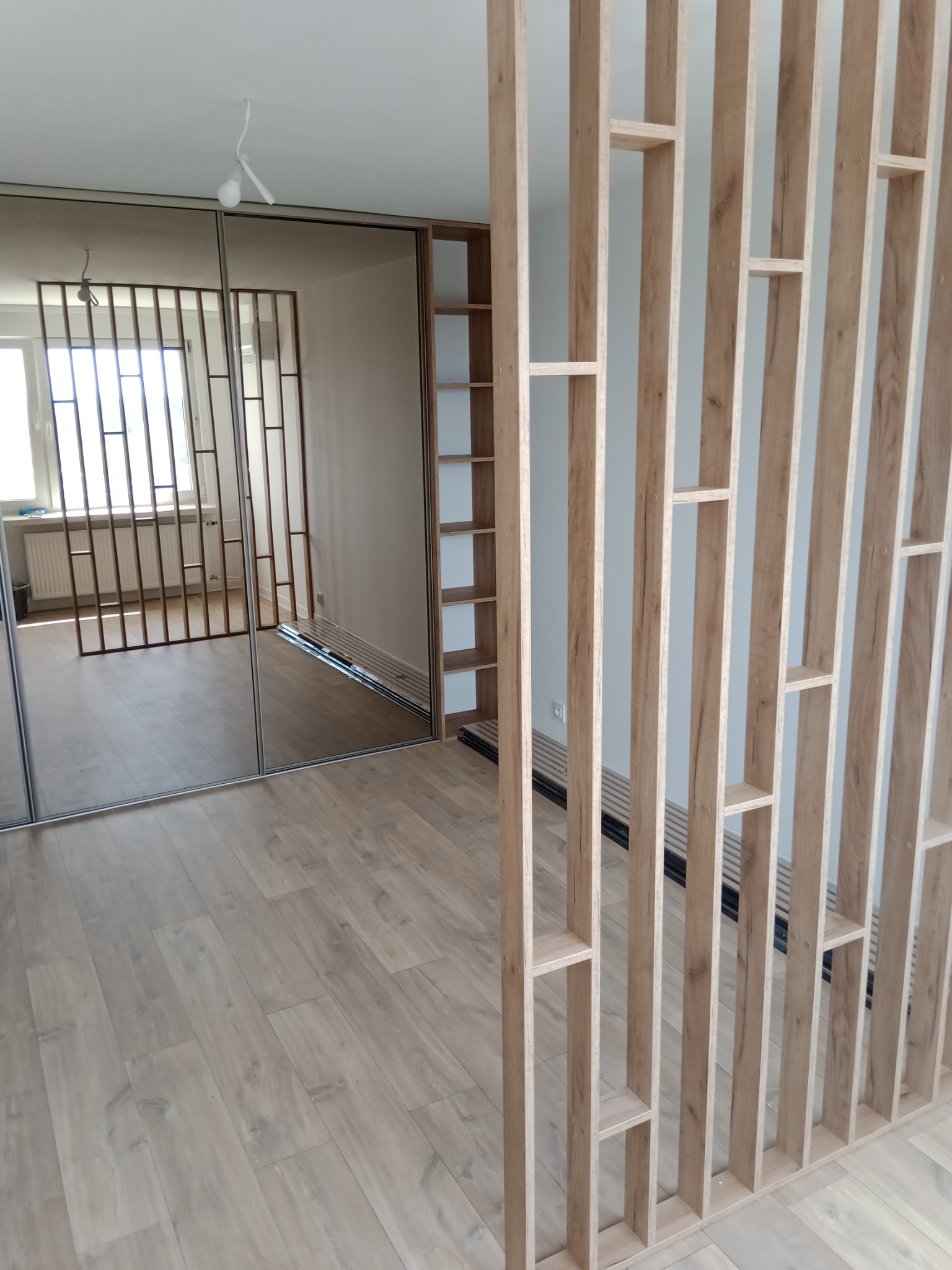 Nowoczesny pokój z drewnianą ażurową ścianką działową i szafą przesuwną z lustrami, widoczne panele podłogowe i niedokończone oświetlenie sufitowe.