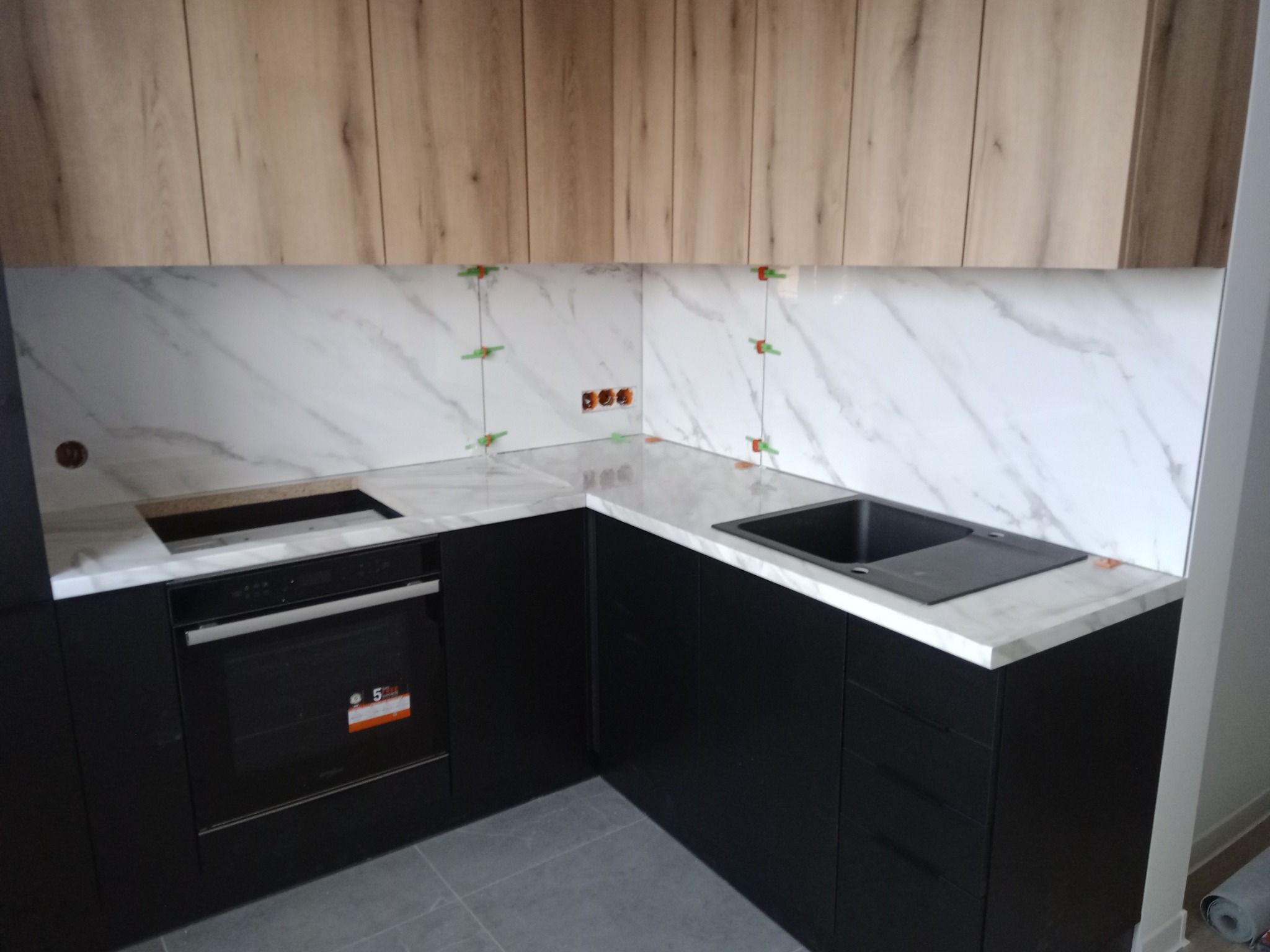 Fragment kuchni w trakcie wykańczania: czarne szafki dolne, marmurowy blat i panel ścienny z dystansami, drewnopodobne szafki górne, widoczne gniazdka elektryczne.