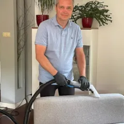 Mężczyzna w czarnych rękawicach czyści narożnik sofy za pomocą profesjonalnego odkurzacza piorącego z przezroczystą końcówką, w tle widoczny biały regał i doniczki z roślinami.