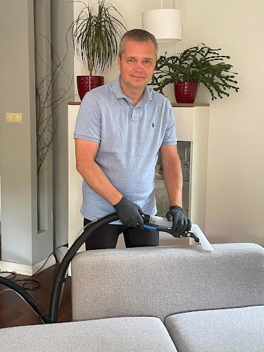 Mężczyzna w czarnych rękawicach czyści narożnik sofy za pomocą profesjonalnego odkurzacza piorącego z przezroczystą końcówką, w tle widoczny biały regał i doniczki z roślinami.