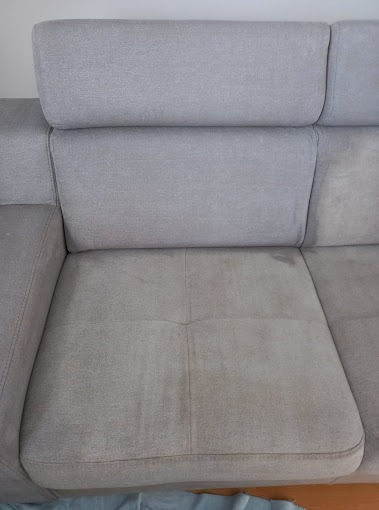 Fragment szarej, tekstylnej kanapy z widocznymi plamami na siedzisku przed czyszczeniem.