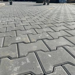DipBud - Szara, betonowa kostka brukowa ułożona w jodełkę. Widoczne fugi i krawężnik. W tle fragmenty palet i sylwetki dwóch osób.