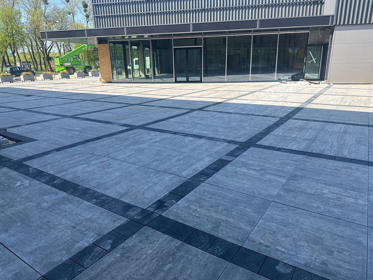 Szara kostka brukowa ułożona w geometryczny wzór przed nowoczesnym budynkiem z przeszkloną fasadą; widoczny podnośnik koszowy w tle.