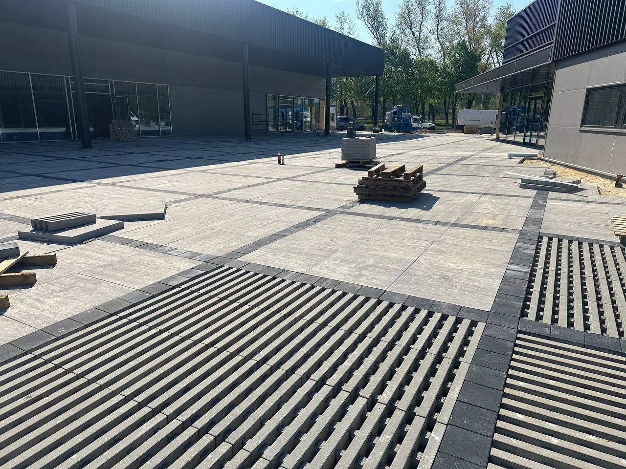 Szara kostka brukowa z czarnymi obrzeżami i kratkami odpływowymi na dużej powierzchni placu przed budynkiem, palety z kostką w tle.