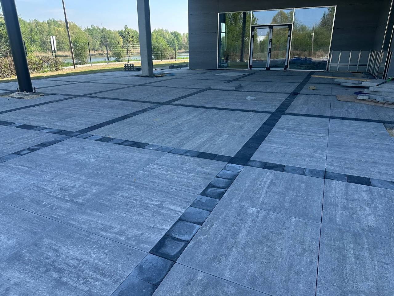 Szara kostka brukowa ułożona w geometryczny wzór z ciemnymi obramowaniami, widoczne fragmenty budynku w tle.