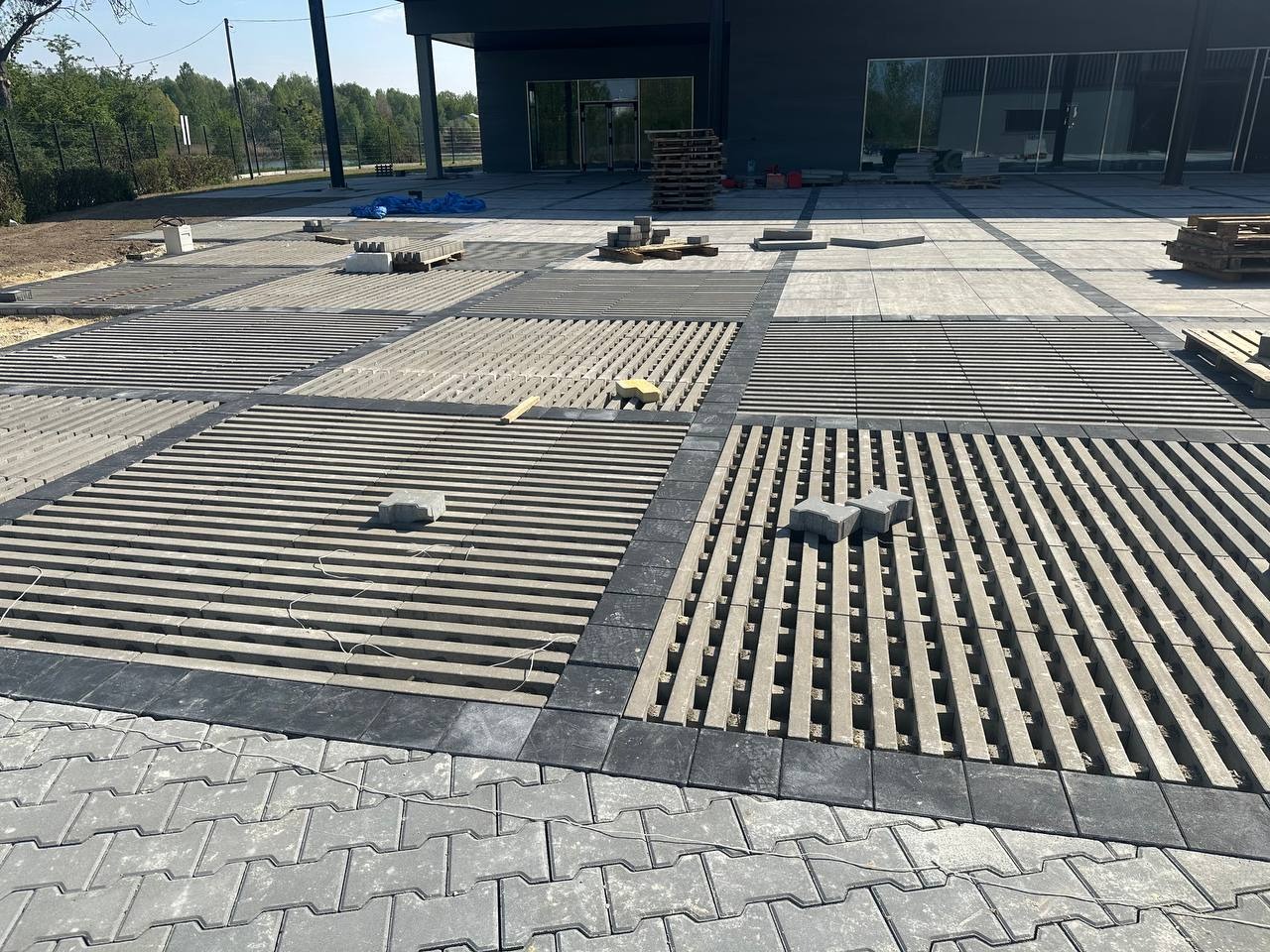 Etap prac: przygotowanie podłoża pod kostkę brukową w Katowicach. Widoczne ułożone rzędy z przerwami, palety z kostką, budynek w tle.