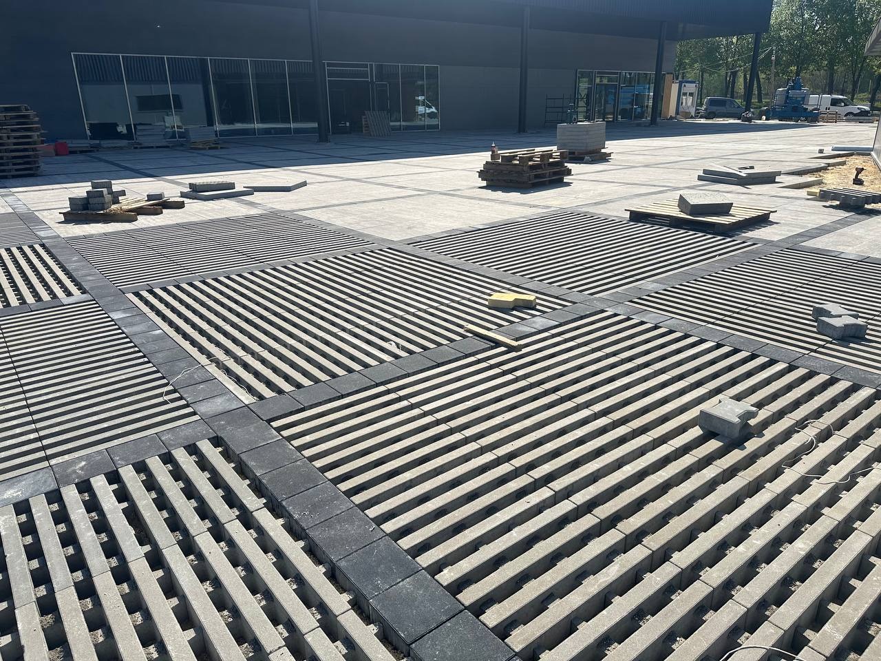 Układanie ażurowej kostki brukowej w geometryczny wzór na placu. Widoczne palety z kostką i częściowo ułożone fragmenty nawierzchni. Nowoczesny design.