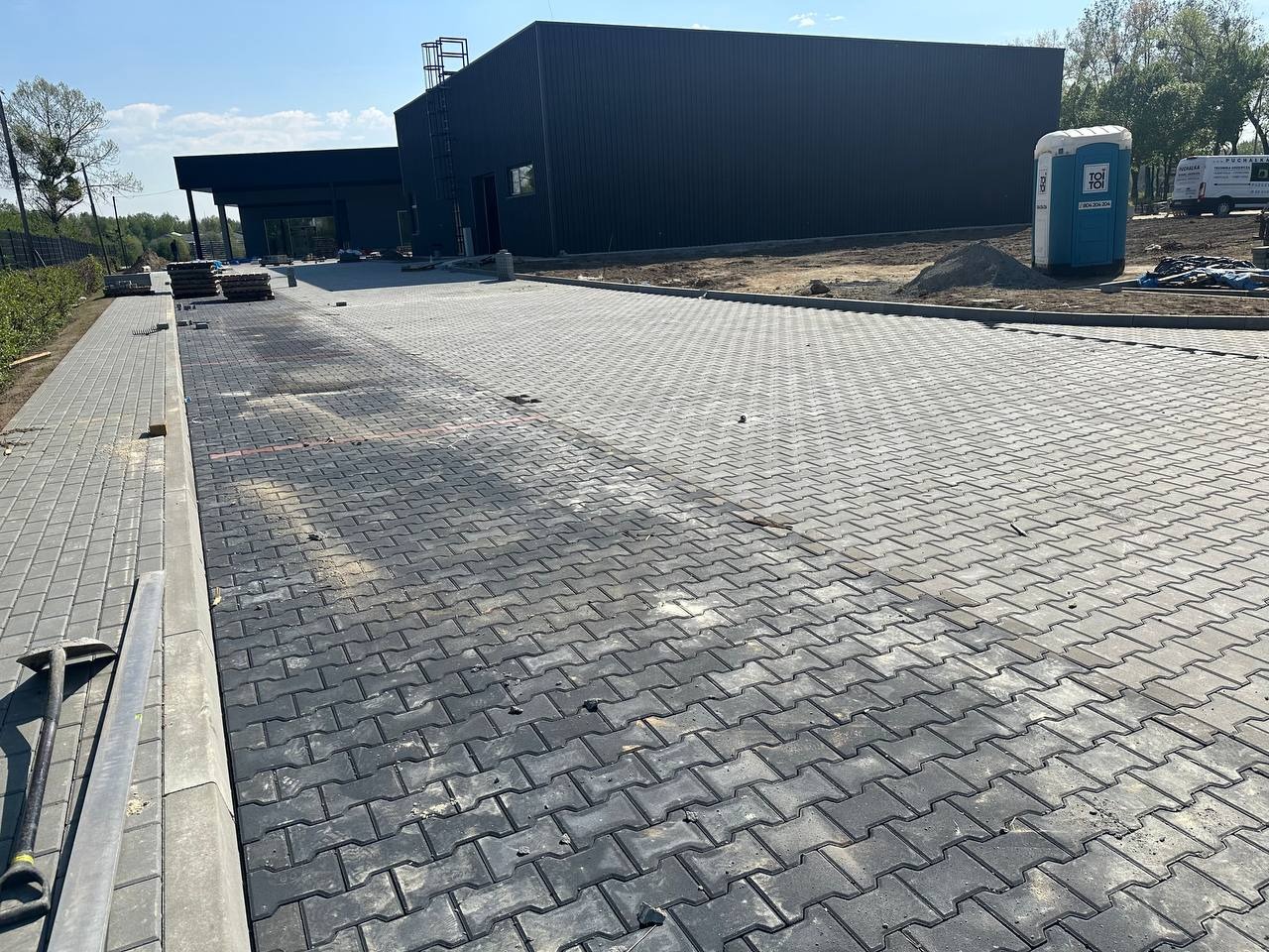 Częściowo ułożona, szara kostka brukowa na rozległym terenie budowy, z widocznymi narzędziami i budynkiem w tle, w słoneczny dzień.