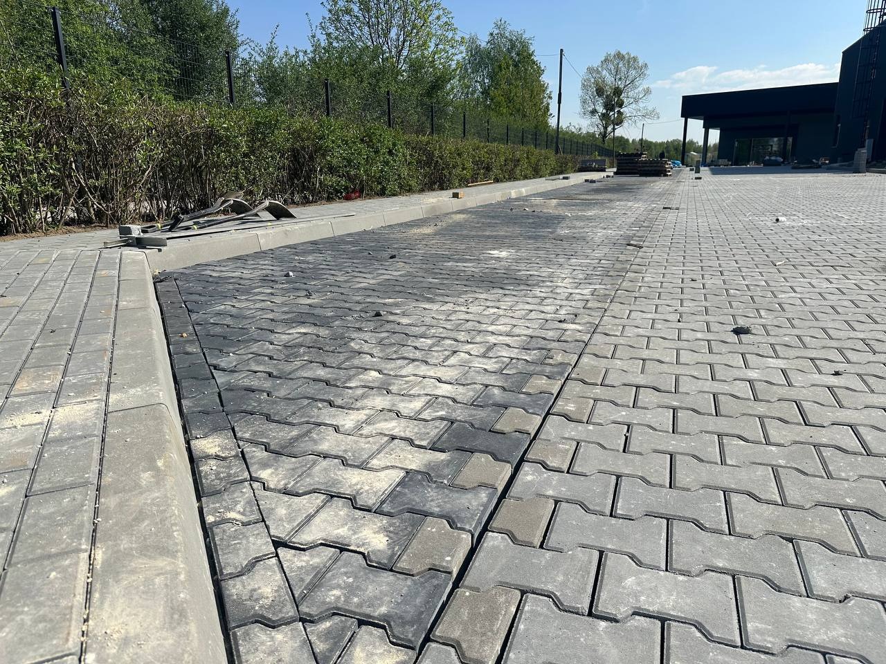 Częściowo ułożona, szara kostka brukowa typu I na podjeździe, z widocznymi narzędziami i elementami podsypki, otoczona zielenią i budynkami w tle.