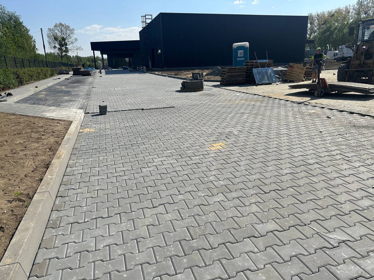 Szara kostka brukowa ułożona we wzór jodełki na podjeździe przed budynkiem. Widoczne palety z kostką i sprzęt budowlany. Częściowo ułożona kostka.