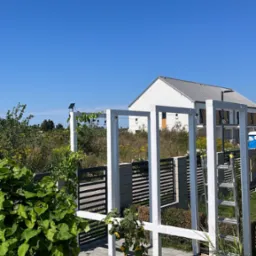 Biała pergola z oświetleniem solarnym, pnączami i wiszącymi żarówkami w ogrodzie z białym żwirem, w tle dom z szarą dachówką i drabina oparta o konstrukcję.