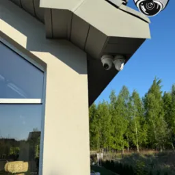 Dwie białe kamery monitoringu kopułkowe zamontowane pod okapem domu, z logo firmy w rogu. Widok na zewnątrz, drzewa i błękitne niebo w tle.