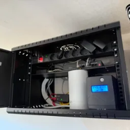 Szafa rack z zainstalowanym sprzętem sieciowym: router TP-Link, zasilacz awaryjny UPS, listwa zasilająca Lanberg i switch Dahua. Widoczne kable. Otwarta szafa w kolorze czarnym.