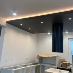 Wnętrze kuchni w trakcie montażu: szare szafki bez frontów, czarny okrągły okap, podwieszany sufit z oświetleniem LED, kartony z nowym sprzętem AGD.