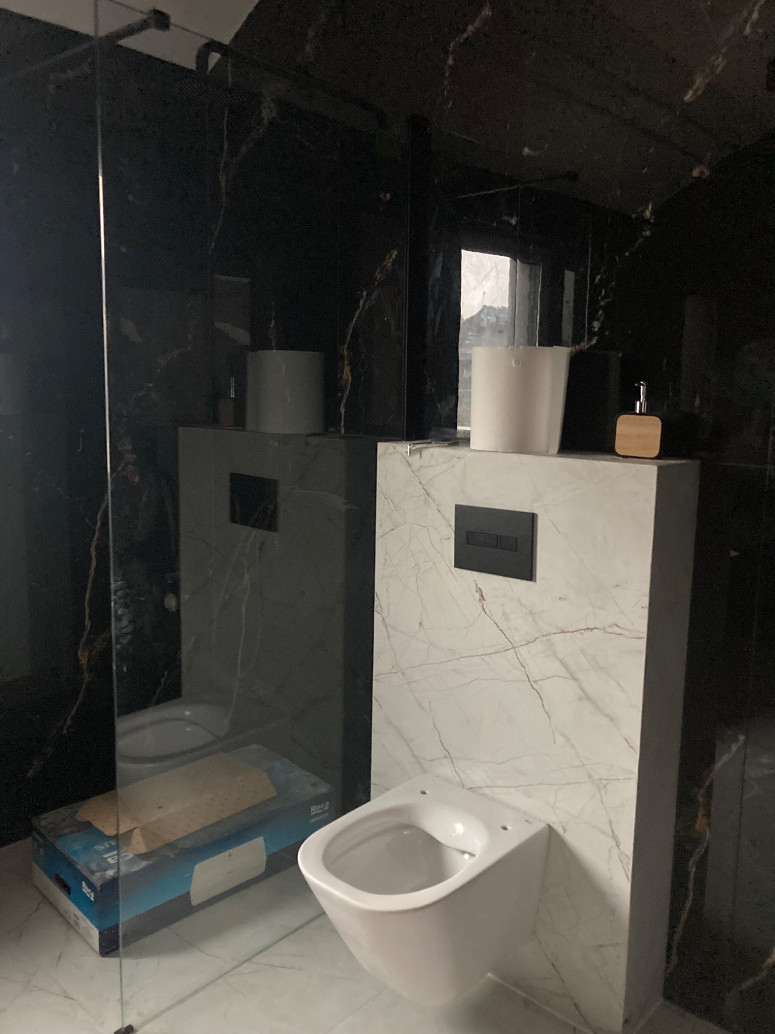 Montaż wiszącej toalety Roca na stelażu podtynkowym z marmurową okładziną w łazience wykończonej czarnym marmurem, widoczna szklana kabina prysznicowa.