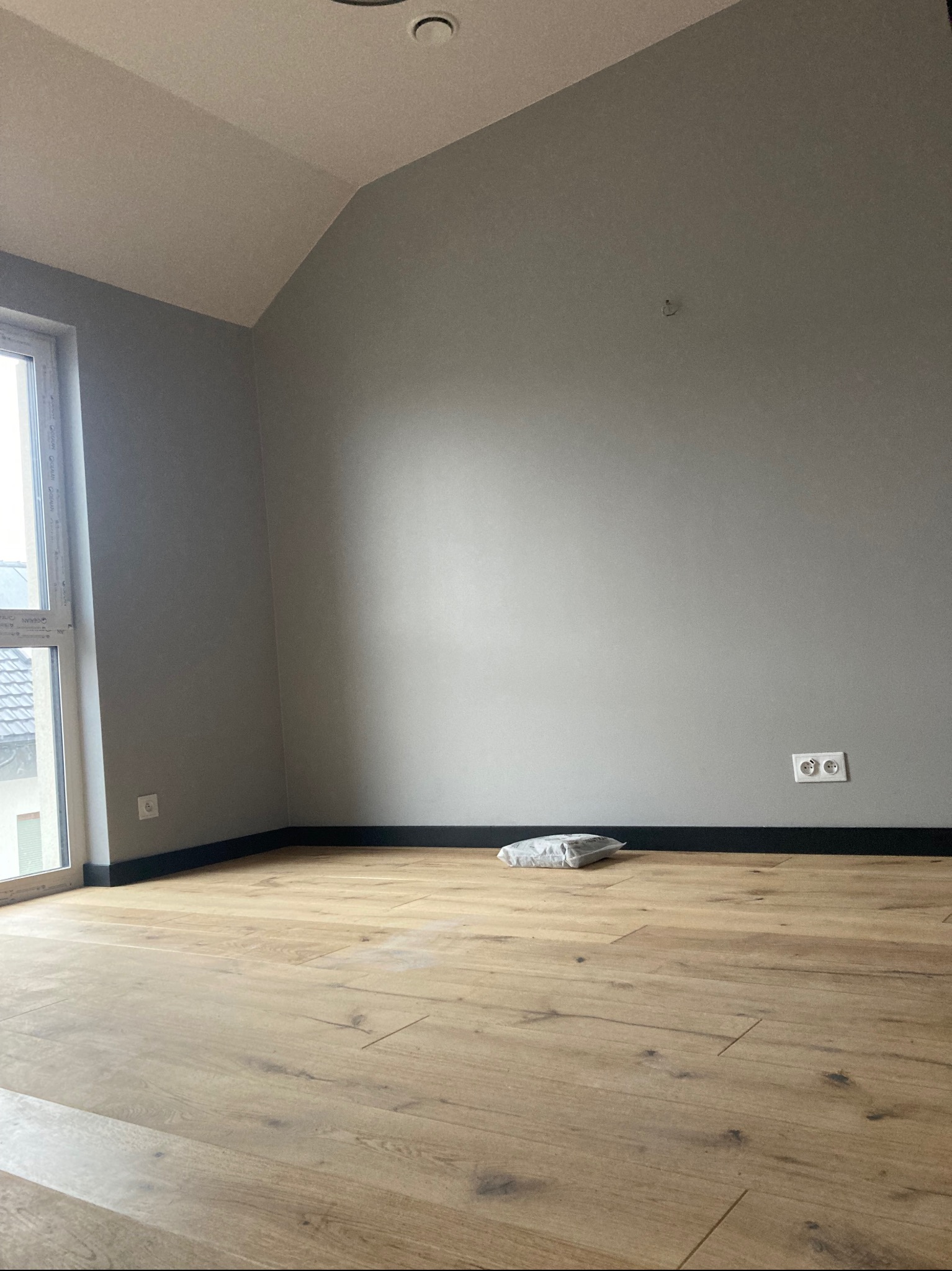 Pokój po remoncie: świeżo pomalowane szare ściany, ułożona jasna podłoga z paneli, widoczne listwy przypodłogowe i gniazdka elektryczne, na podłodze leży paczka z materiałami.