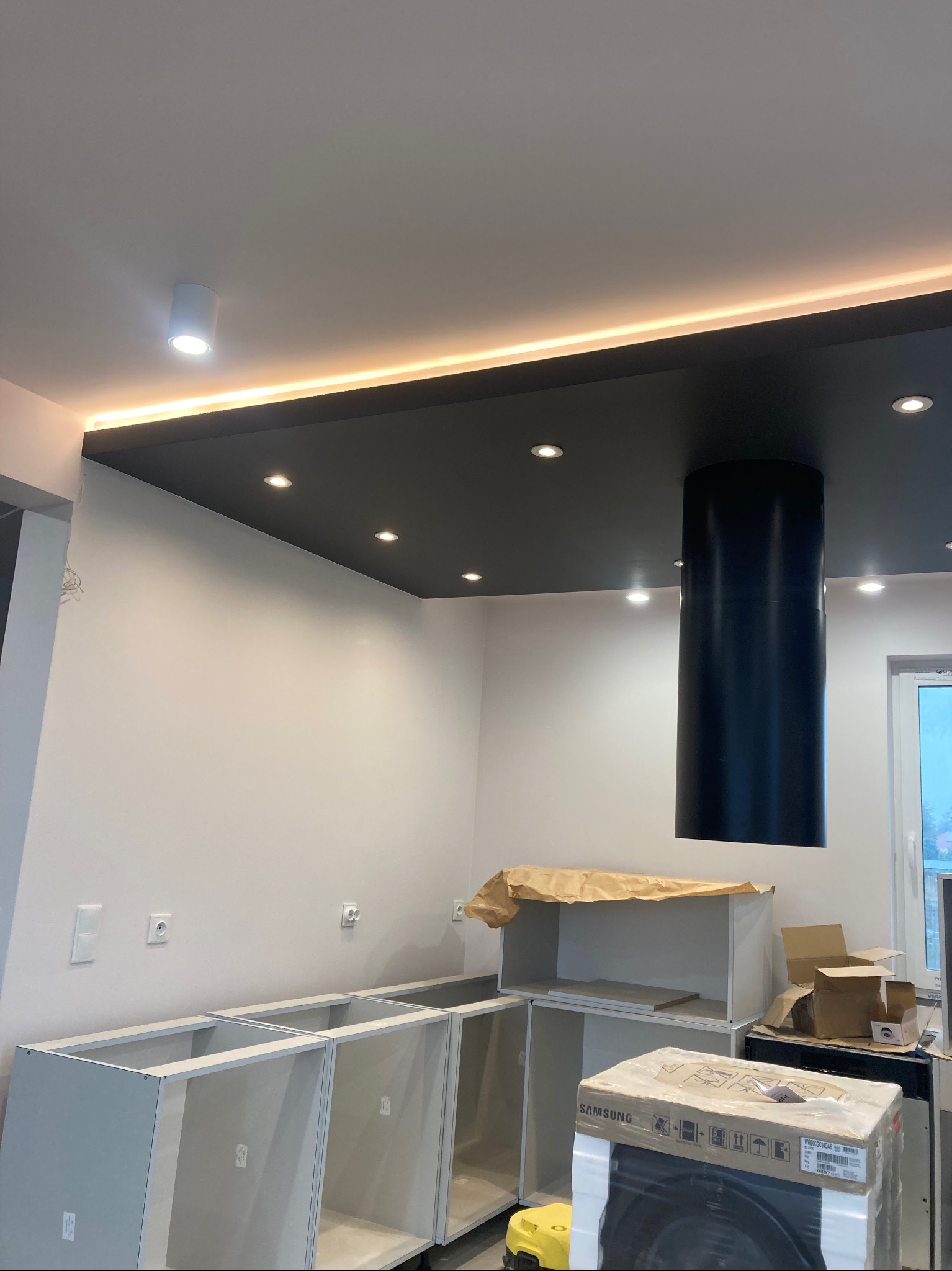 Wnętrze kuchni w trakcie montażu: szare szafki bez frontów, czarny okrągły okap, podwieszany sufit z oświetleniem LED, kartony z nowym sprzętem AGD.