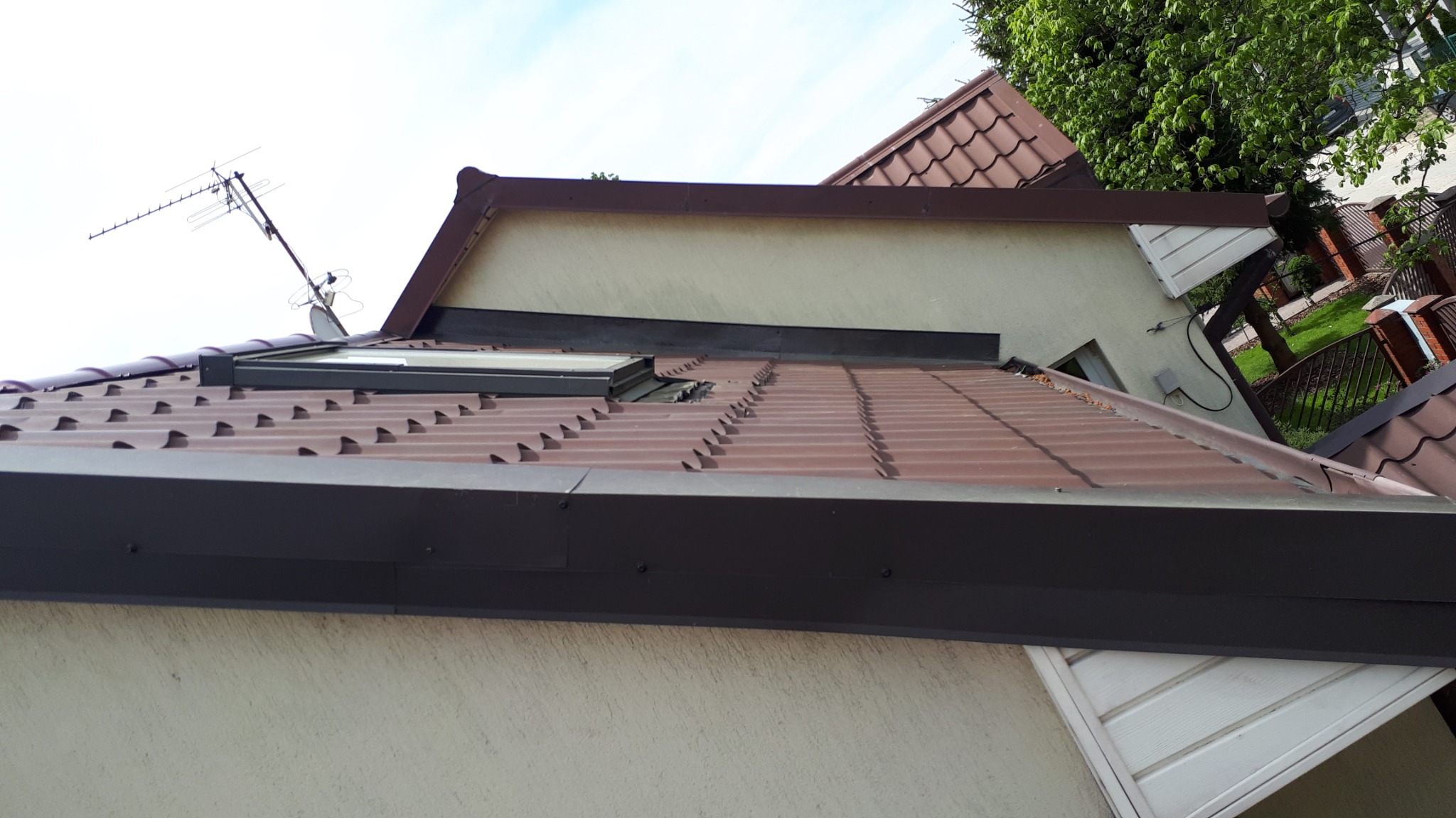 Brązowy dach z blachy trapezowej z oknem dachowym i anteną telewizyjną na tle jasnego nieba i zieleni drzew.