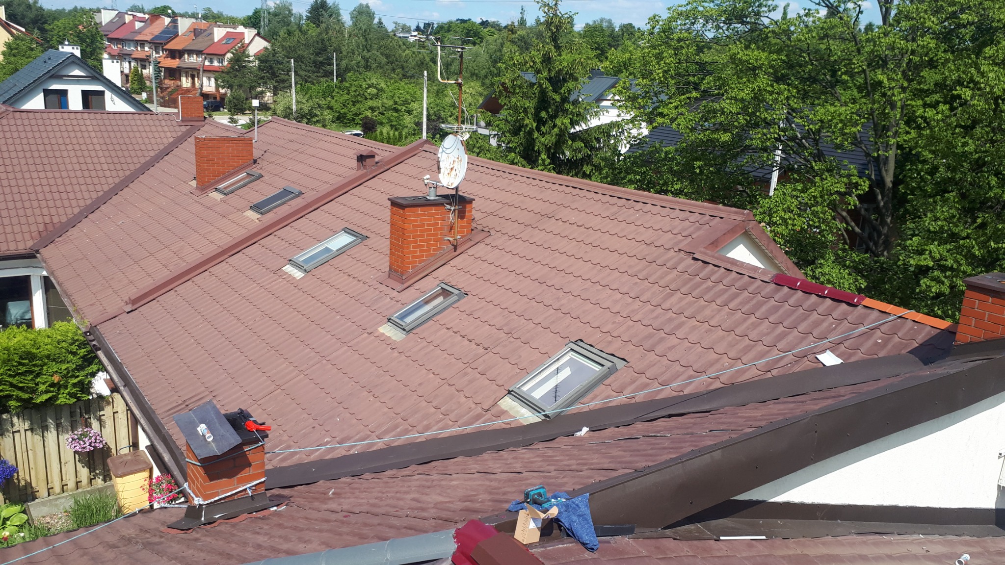 Czerwono-brązowy dach z dachówki ceramicznej z oknami dachowymi i kominami z cegły, na jednym z kominów zamontowana antena satelitarna, narzędzia i materiały budowlane na dachu w trakcie prac.