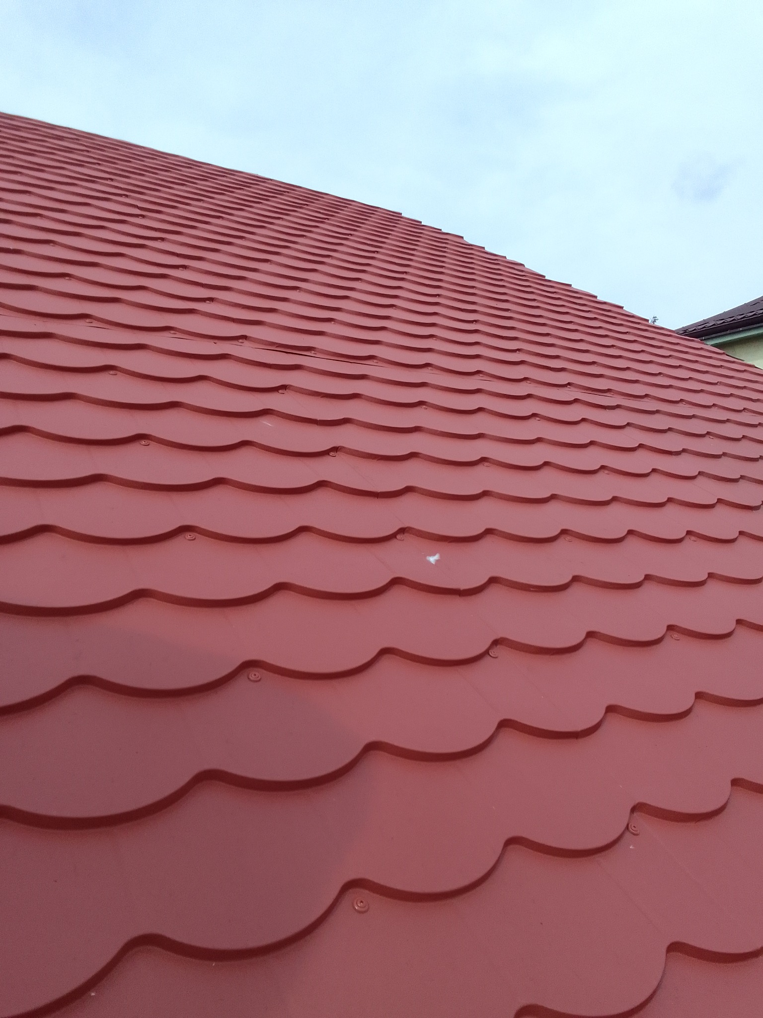 Czerwona blachodachówka modułowa na dachu domu, widoczne mocowania śrubowe, pochmurne niebo w tle.