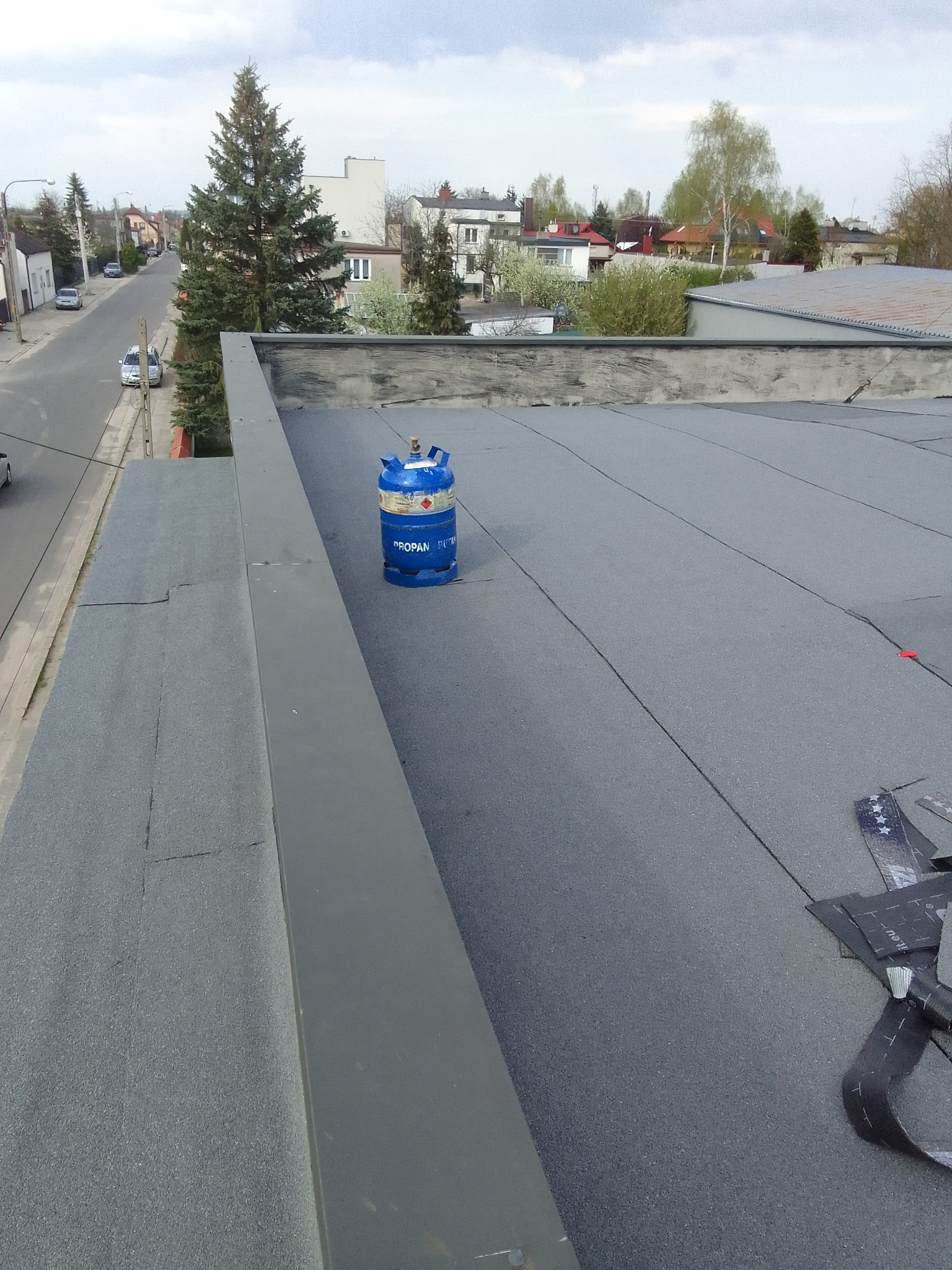 Niebieska butla z propanem na płaskim dachu pokrytym papą, widok z góry na okoliczne budynki i ulicę.